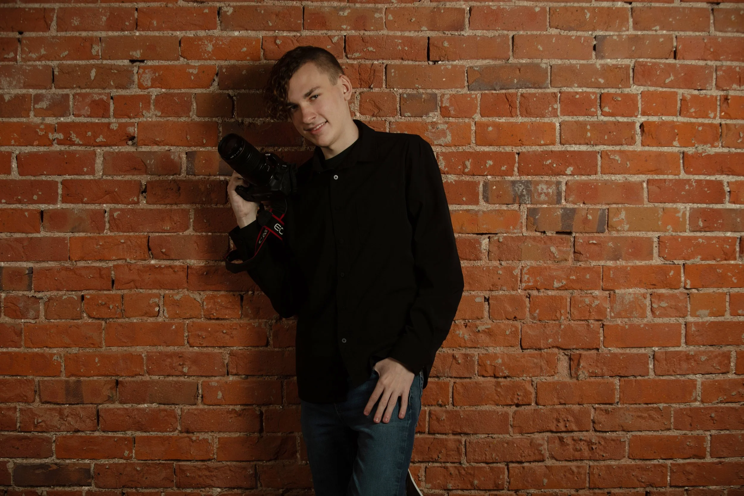A young man with curly hair styled to the side, wearing a black button-up shirt, standing against a red brick wall, holding a camera and smiling.