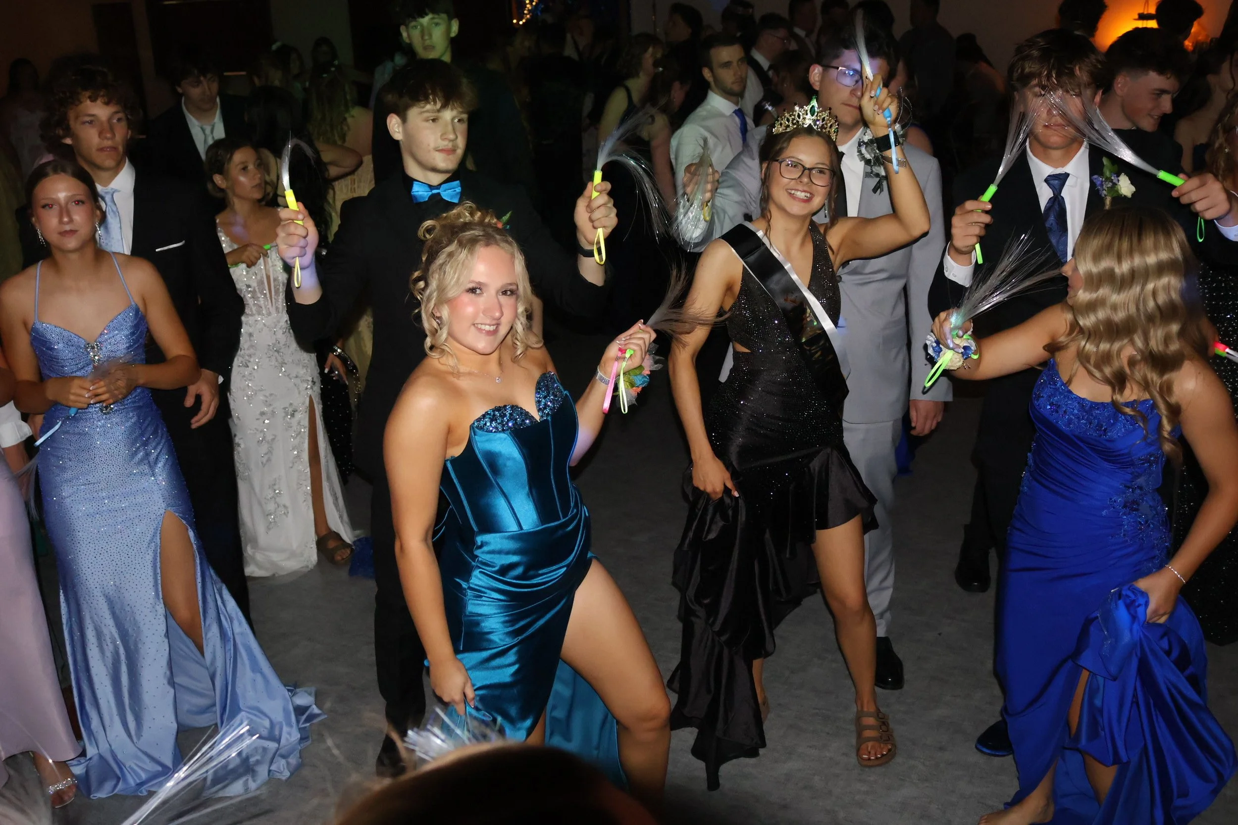 Young people dancing at a formal event, dressed in elegant gowns and suits, with some holding glow sticks and wearing tiaras or crowns.