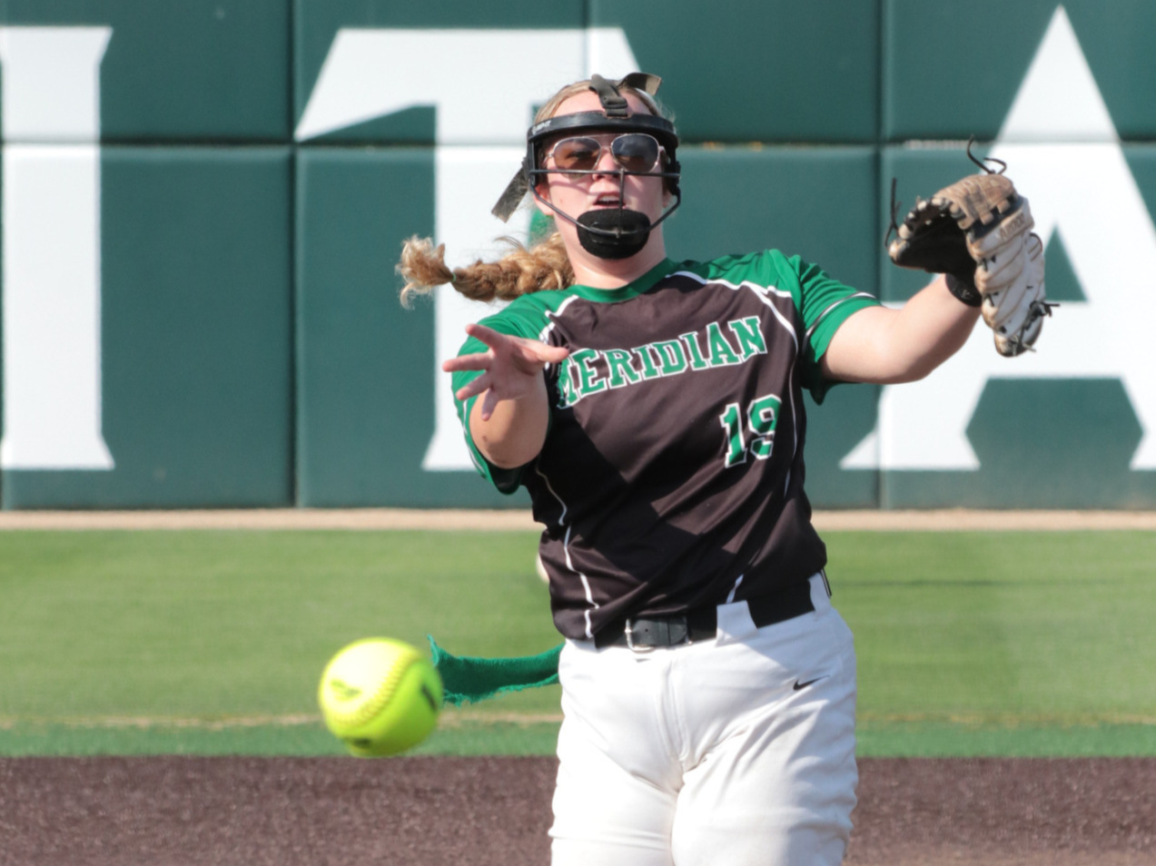 A female softball player in a black and green uniform with 'MERIDIAN' written on it and the number 19, wearing a helmet with a faceguard and sunglasses, reaching out for a yellow softball during a game.
