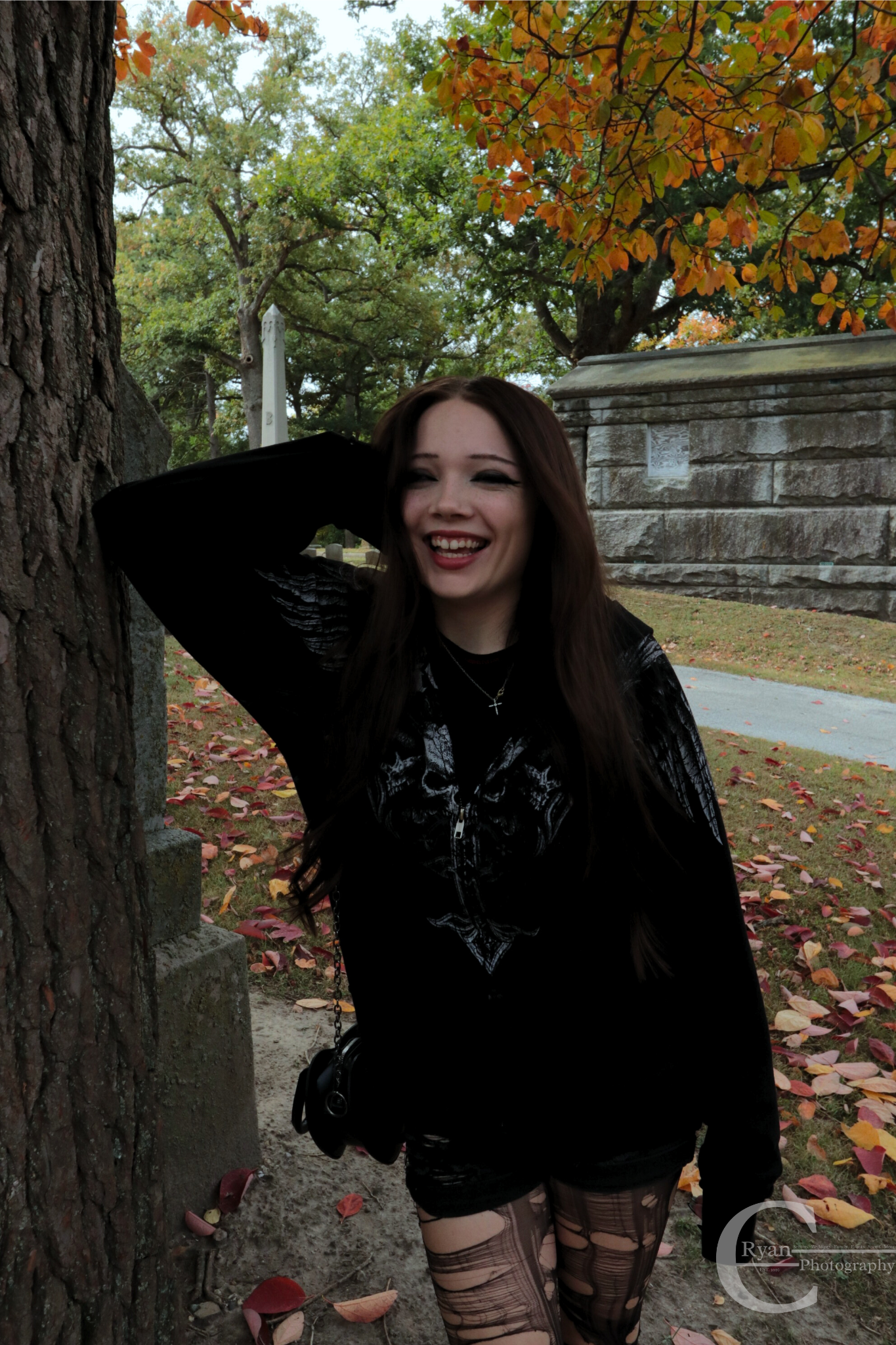 A woman with long dark hair, wearing a black jacket and patterned tights, standing outdoors near a tree with orange and green leaves in the fall. She is smiling and has her hand behind her head. Behind her is a stone structure and fallen leaves on the ground.