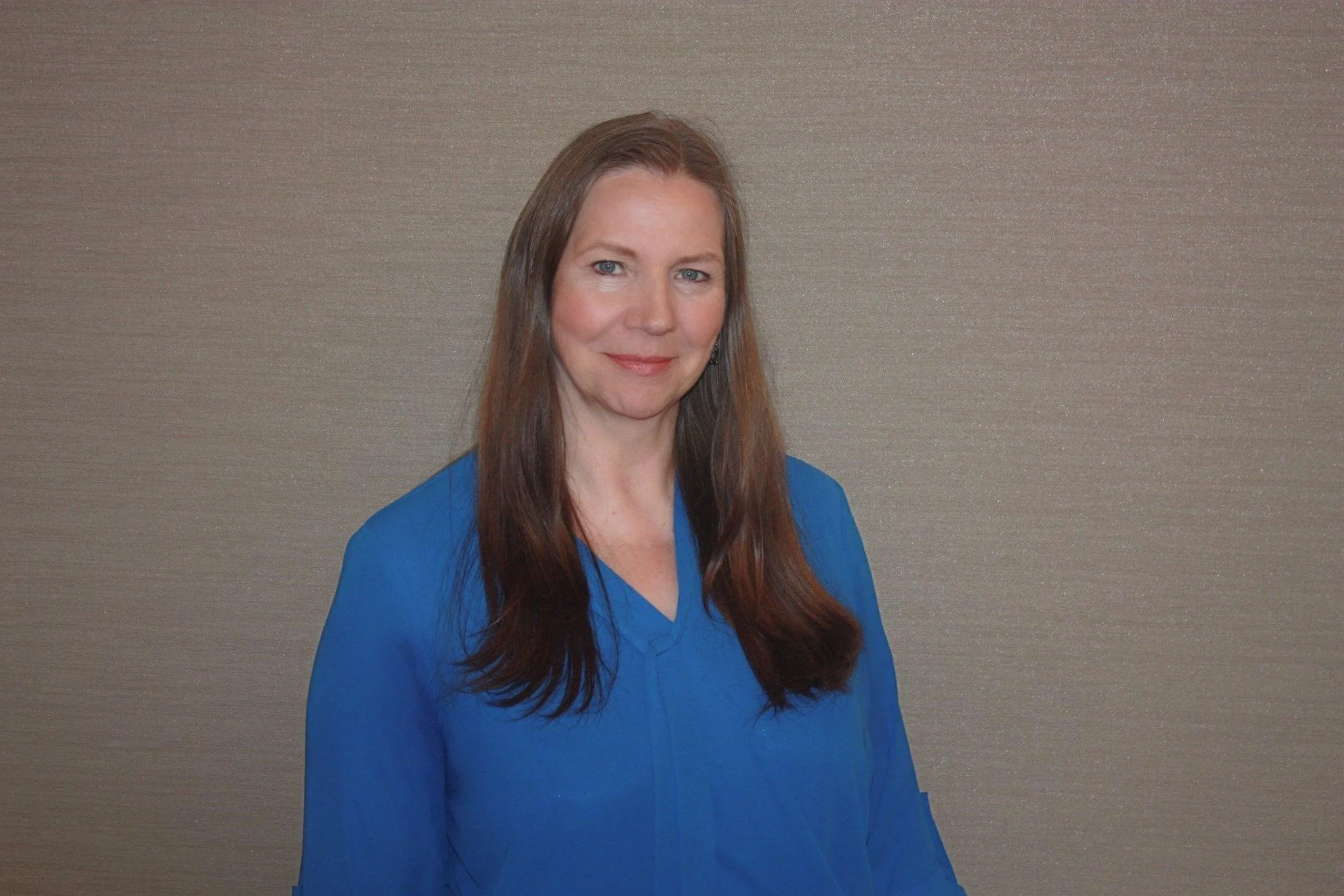 A woman with long brown hair wearing a blue blazer standing against a plain textured wall.