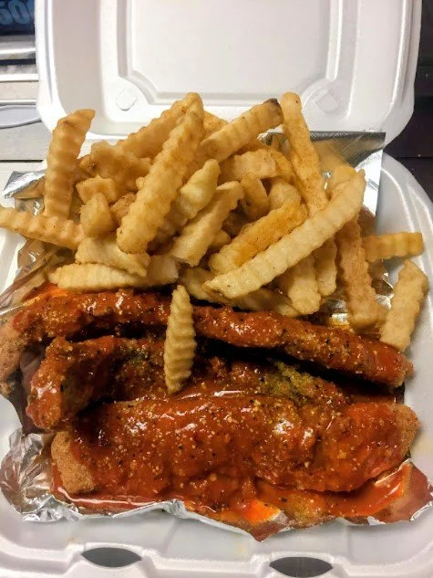 Foil-lined takeout container with fried chicken wings coated in sauce and crinkle-cut French fries.