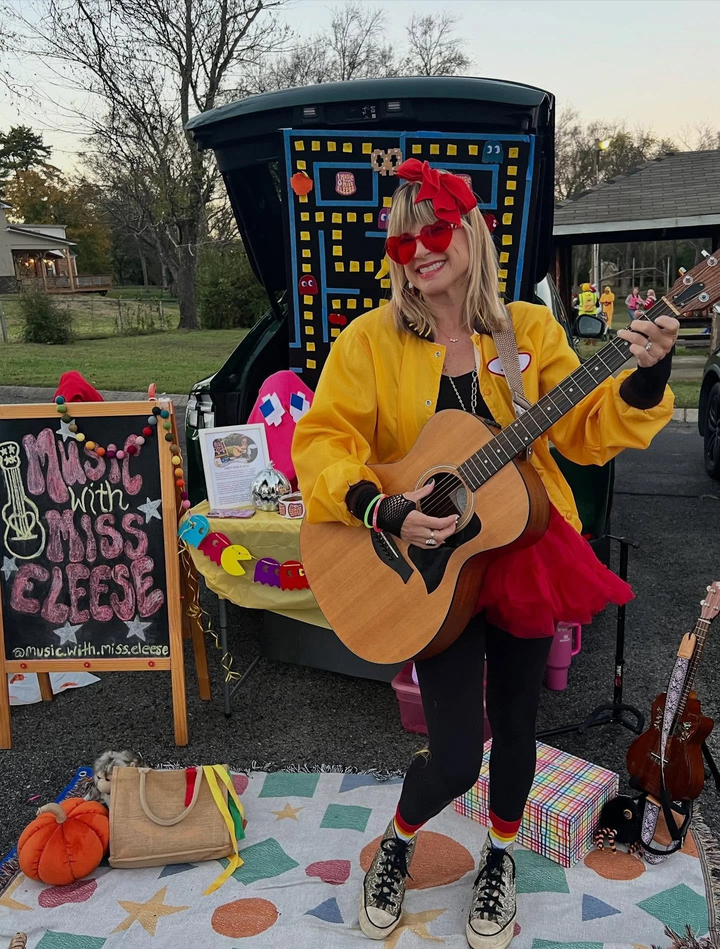 A woman with blonde hair, red sunglasses, and a red bow is playing a guitar and smiling. She is dressed in a yellow jacket, black clothing, colorful socks, and combat boots. She stands in front of a music booth with a sign that reads "Music with Miss Geese" and a decorated Pac-Man-inspired video game backdrop. The booth is outdoors with trees and houses in the background, and there are pumpkins and colorful decorations on the ground.