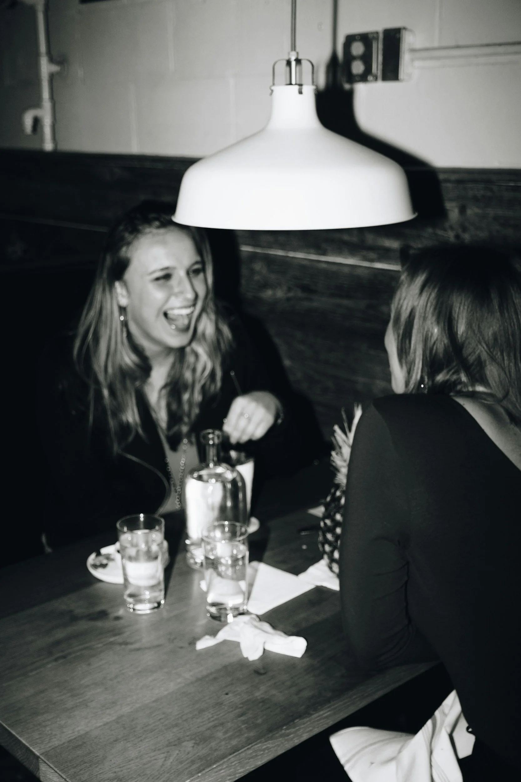 Two women sitting at a table in a restaurant, engaging in conversation and laughing, with drinks in front of them, underneath a hanging light fixture.