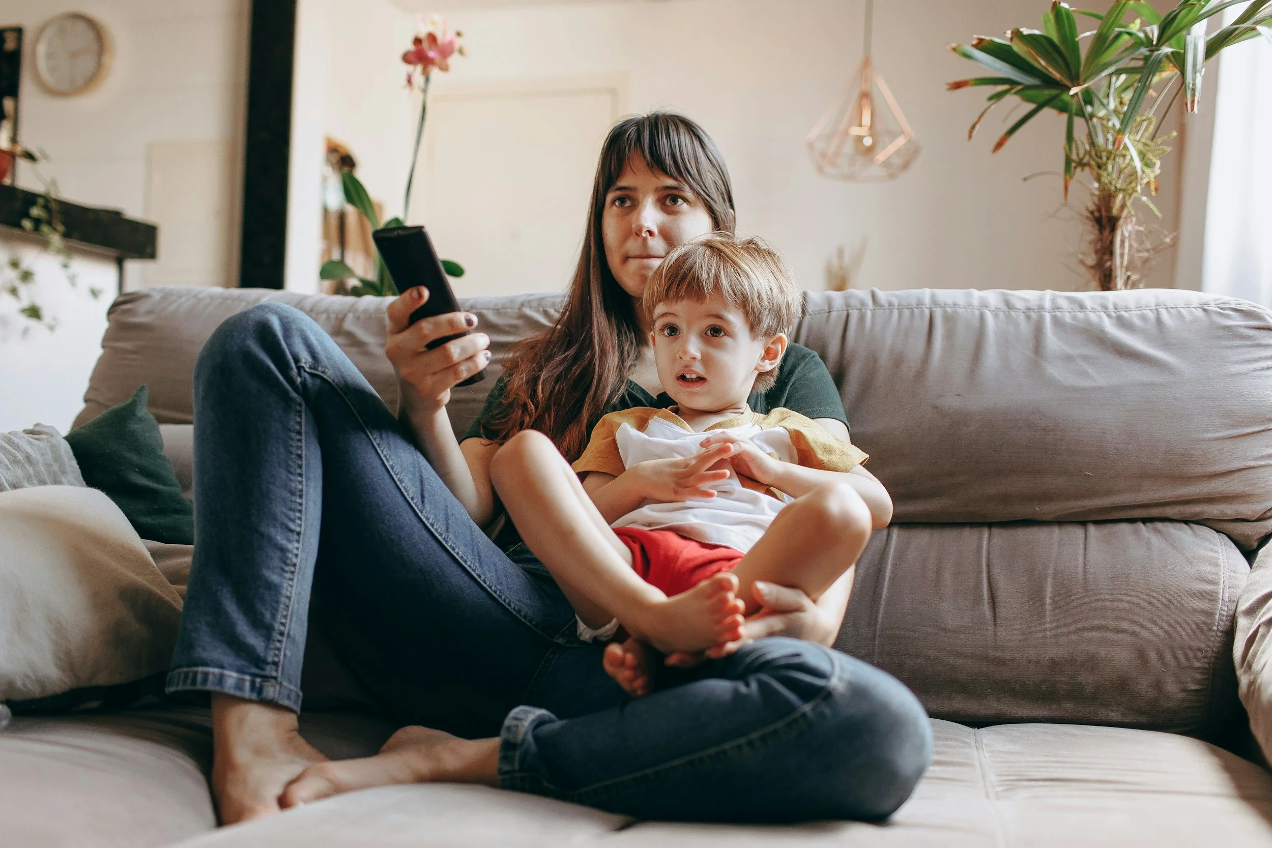 A woman and a young boy sit on a beige couch in a living room, looking at a smartphone. The woman has long dark hair and is wearing a black shirt and blue jeans, while the boy has light brown hair and is dressed in a yellow and white shirt with red shorts. The living room has several plants, a wall clock, and a decorative light fixture.