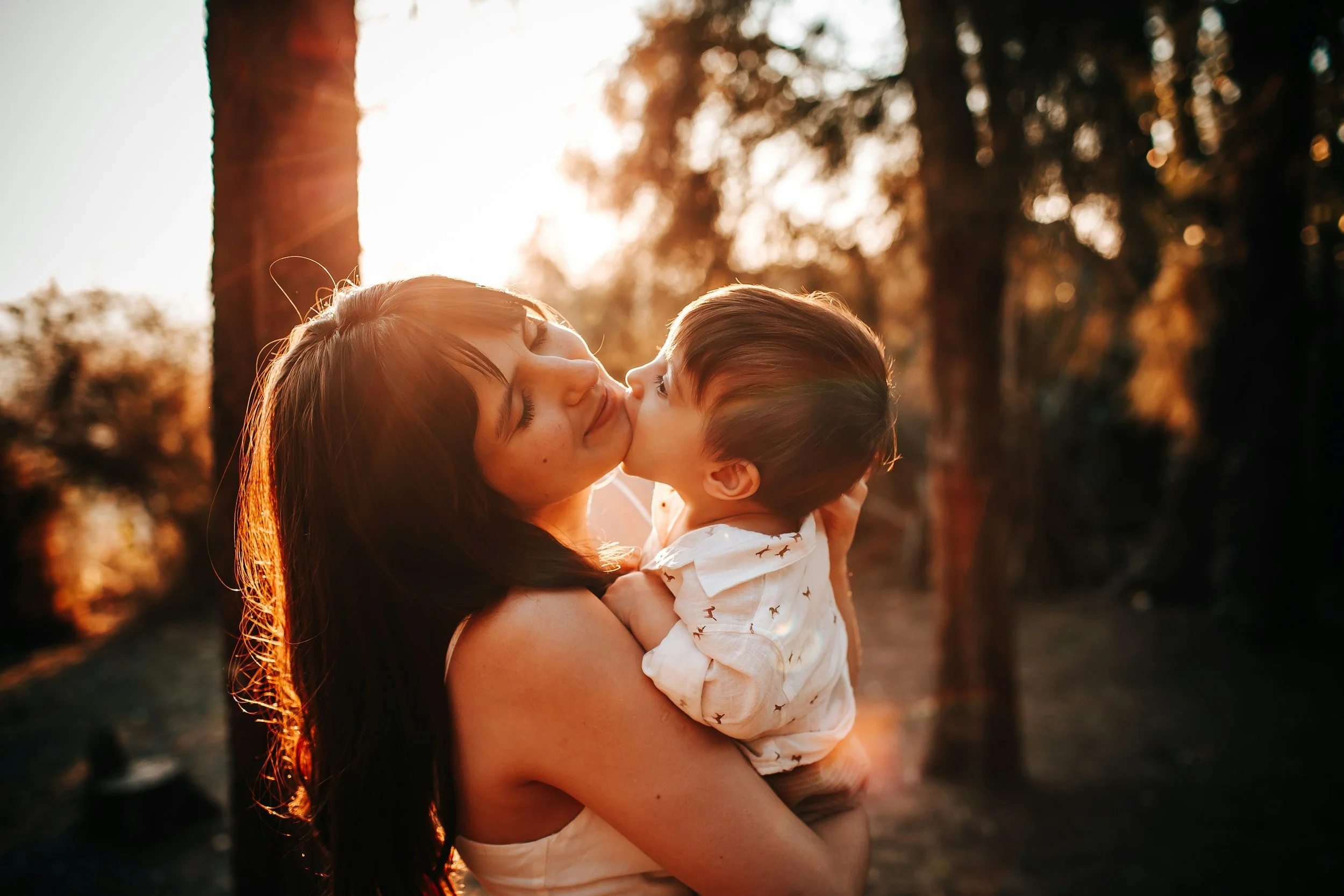 A woman holding a young child outdoors during sunset, with trees and warm sunlight in the background.
