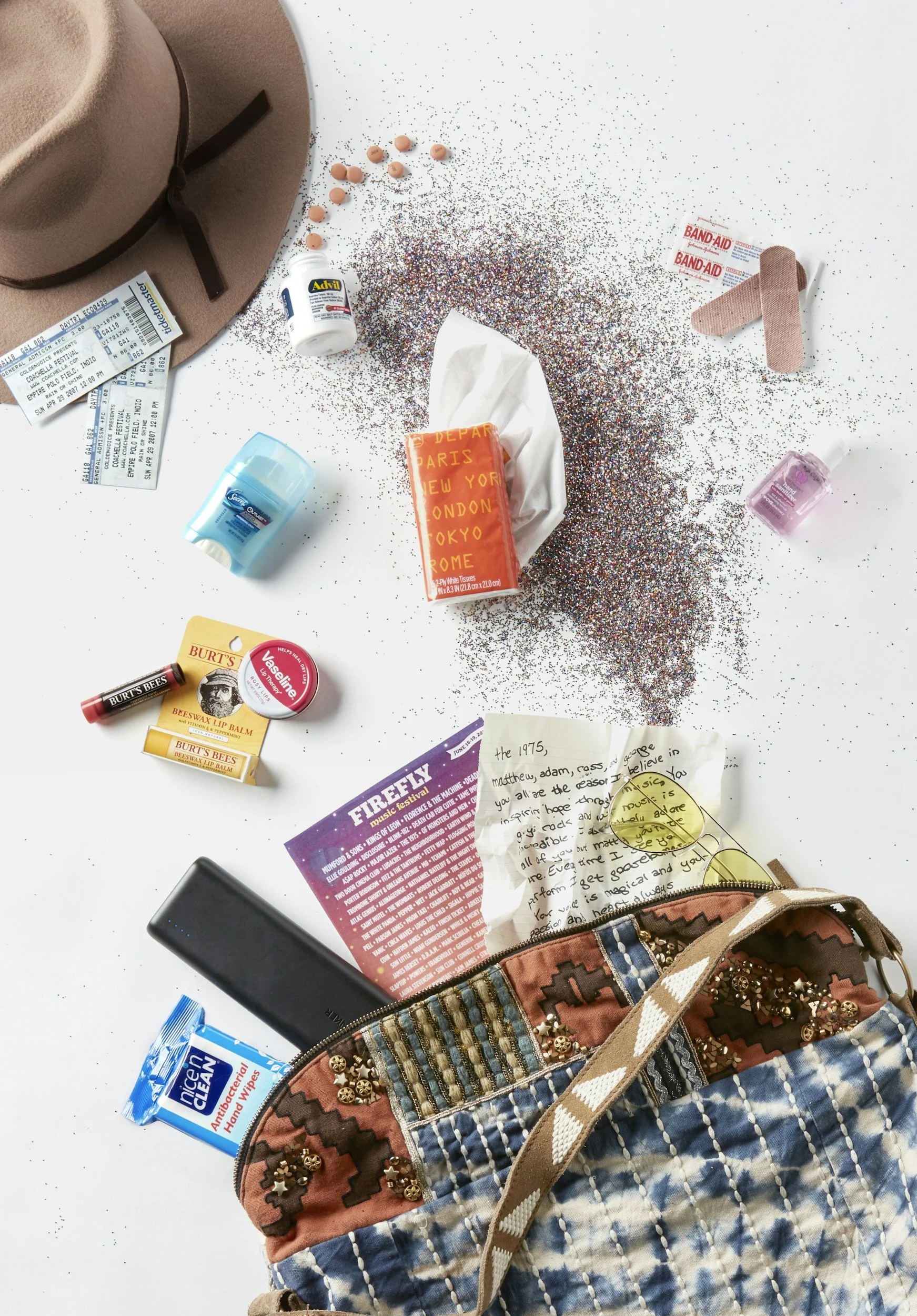 A cluttered white surface with various personal items including a tan hat, tickets, a jar of Advil, a pack of tissues, a mini hand sanitizer, a black device, a packet of antibacterial hand wipes, a handwritten note, and a patterned purse with embellishments.