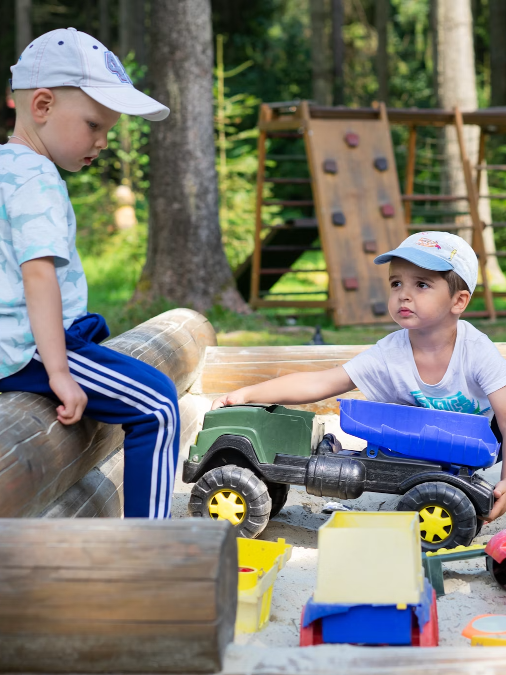 Two young boys playing with toy trucks and sand in an outdoor playground, surrounded by trees and playground equipment.