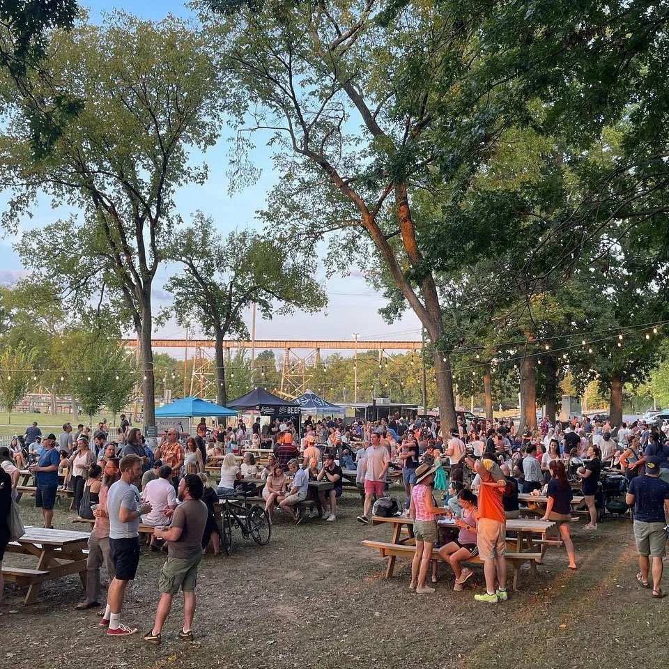 A large outdoor gathering at a park with many people sitting and standing around picnic tables and tents for food and drinks, surrounded by trees with string lights, during evening.
