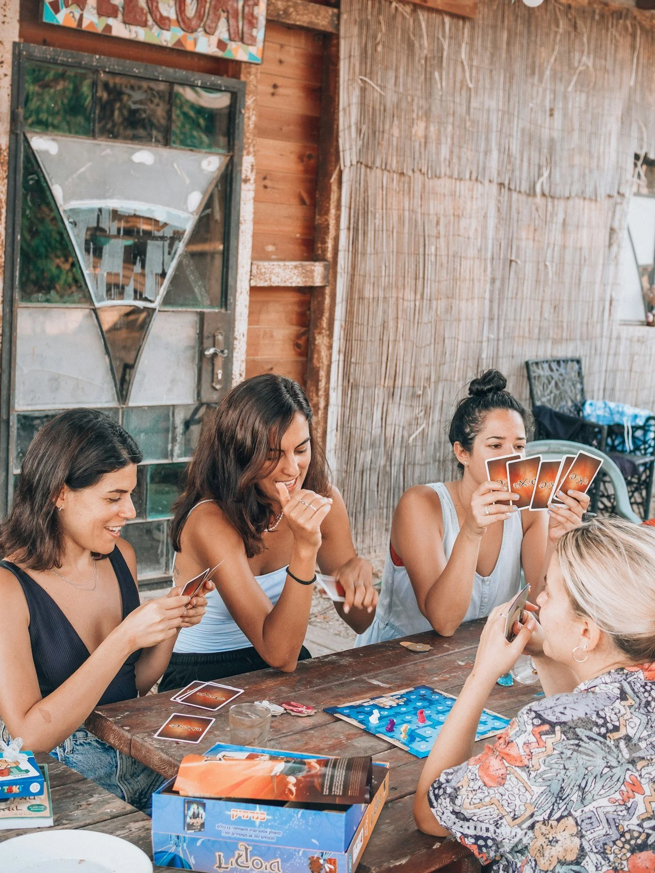 A group of women sitting around a wooden table playing a card game during a gathering or party in a rustic indoor setting with wooden walls and decorative elements.