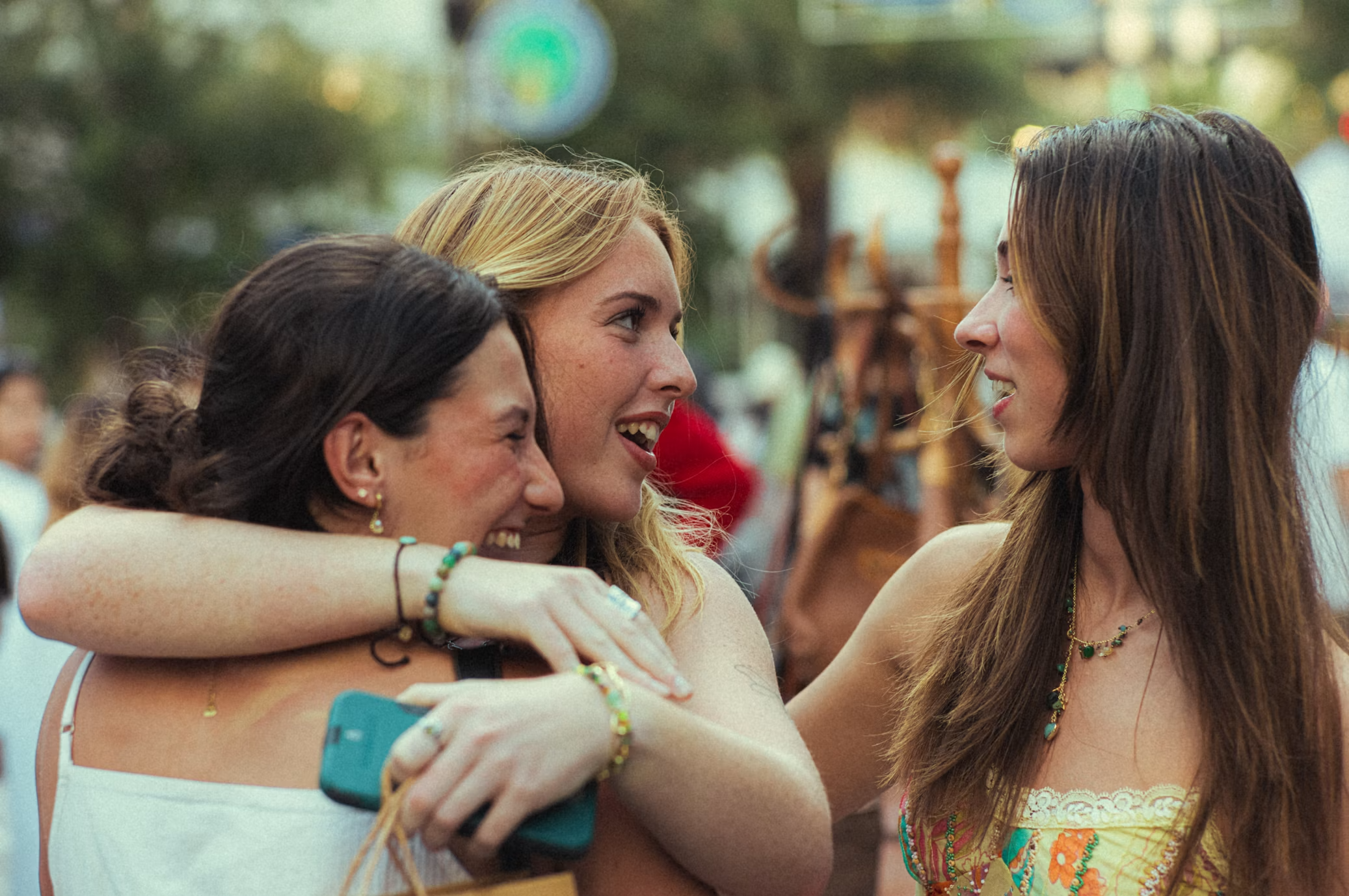 Three women hugging and smiling at each other outdoors at an event, with trees and colorful decorations in the background.