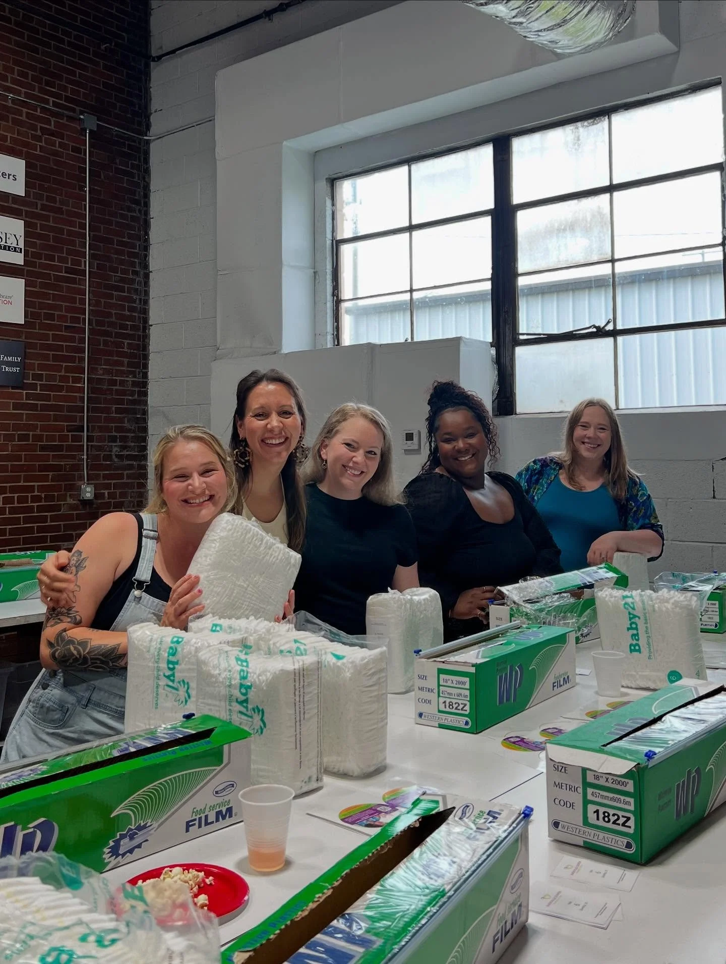 Five women standing behind a table with plastic wrap and boxes of food service film, smiling at the camera inside a room with large window, brick wall, and industrial ceiling.