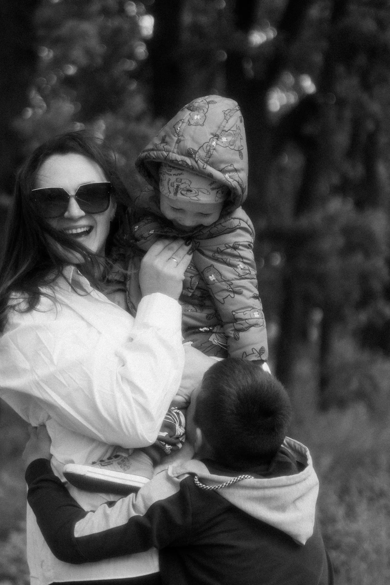 A woman wearing sunglasses smiling and holding a young child wearing a hooded jacket with cartoon characters in an outdoor setting, with blurred trees in the background.