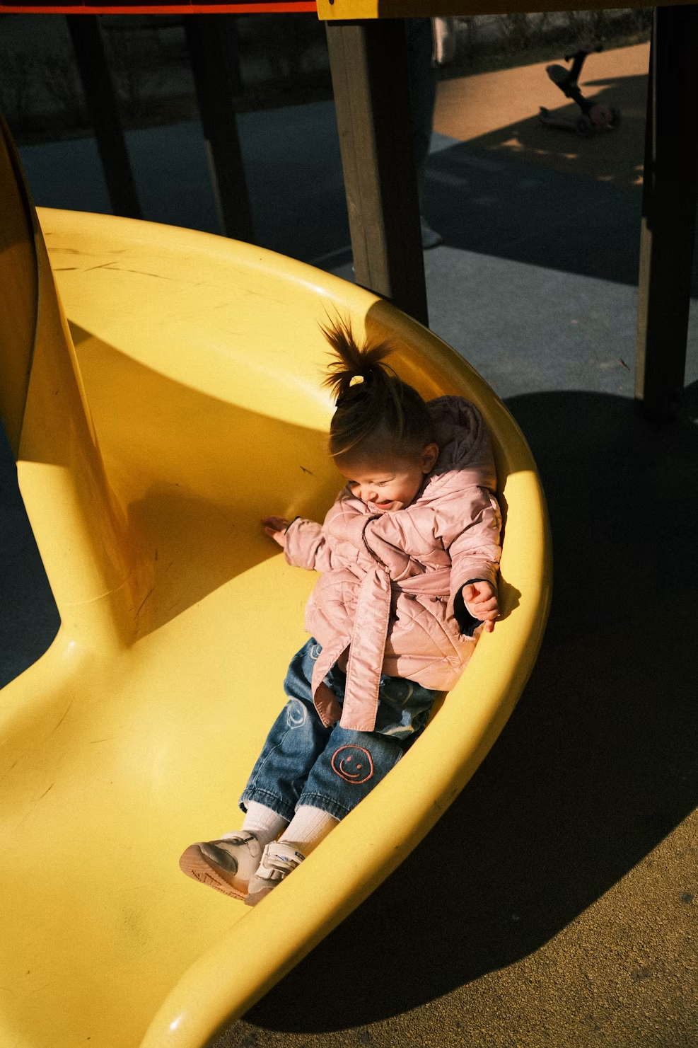 A young girl smiling and playing on a yellow slide at a playground, wearing a pink jacket, jeans, and sneakers, during daytime.