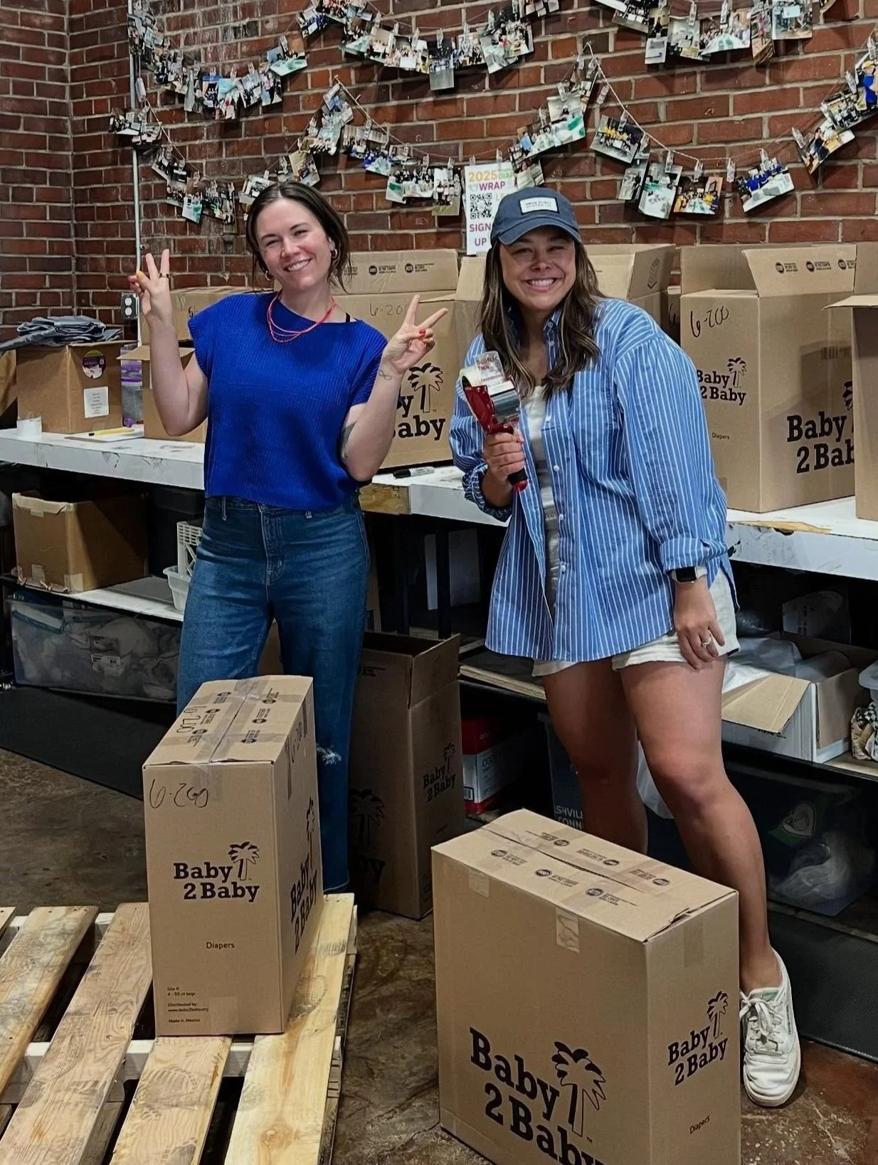 Two women smiling and posing for a photo at a donation or packing event, surrounded by boxes labeled 'Baby 2 Baby' and photos hanging on a brick wall in the background.
