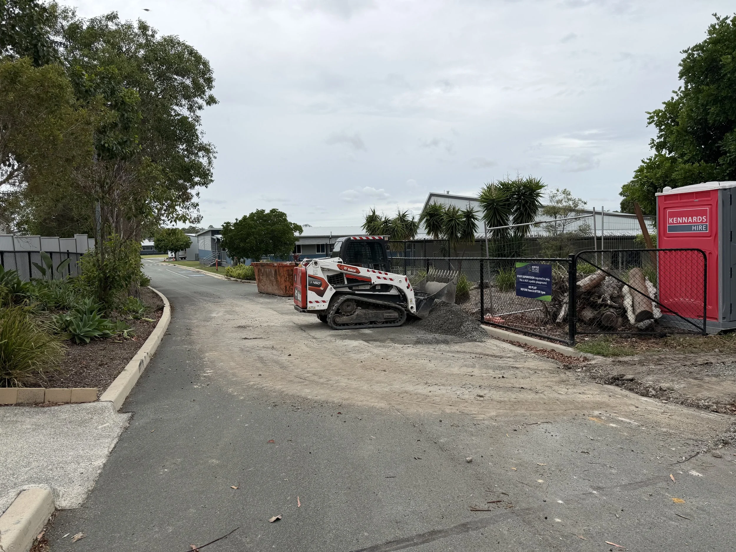 A street under construction with construction equipment, a large orange container, and a red portable toilet. There are trees and residential buildings in the background under a cloudy sky.