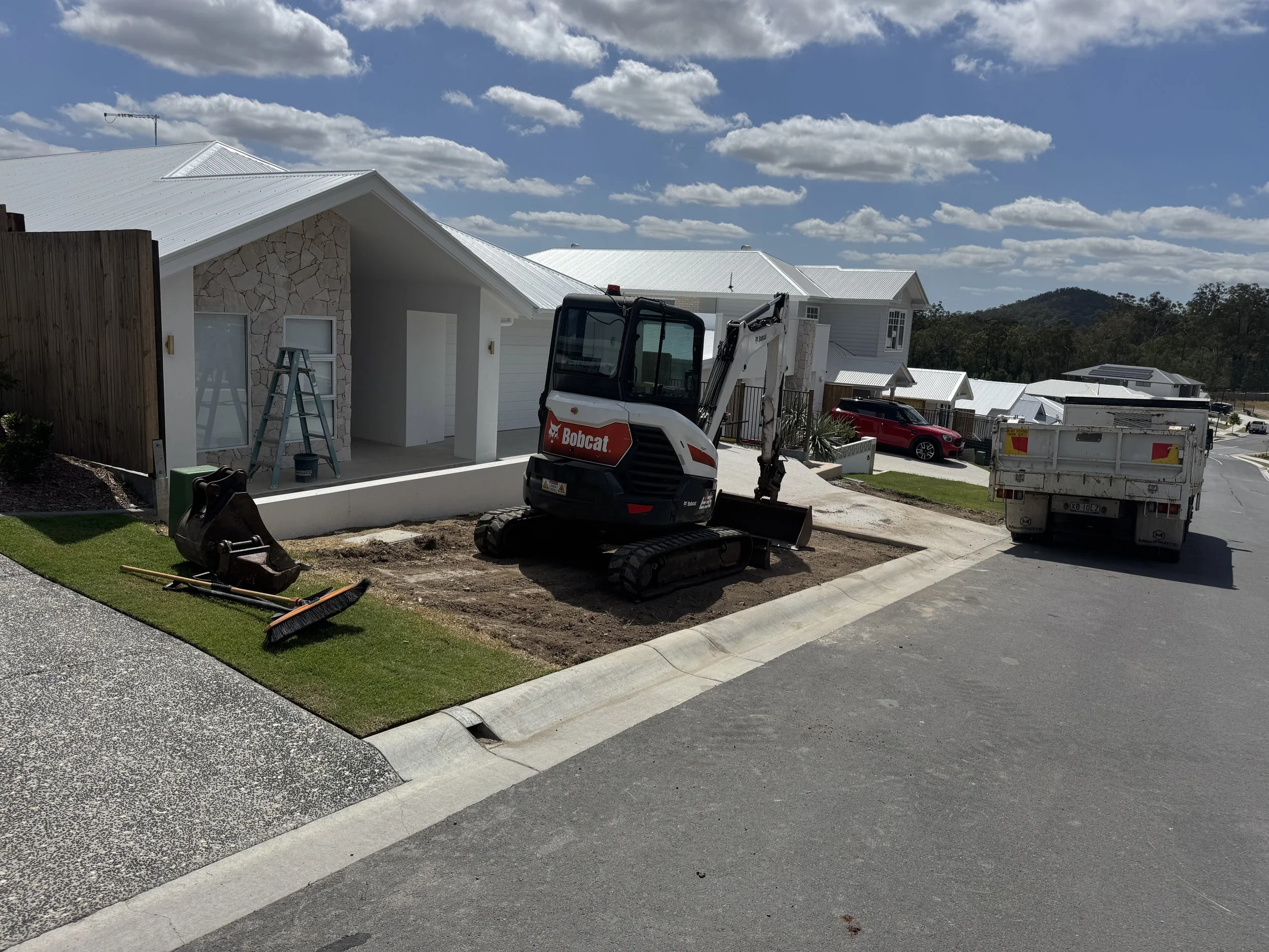 A residential street with houses that have white metal roofs. There is a construction site with a small Bobcat excavator, a ladder, and construction materials. The sky is partly cloudy with scattered clouds.