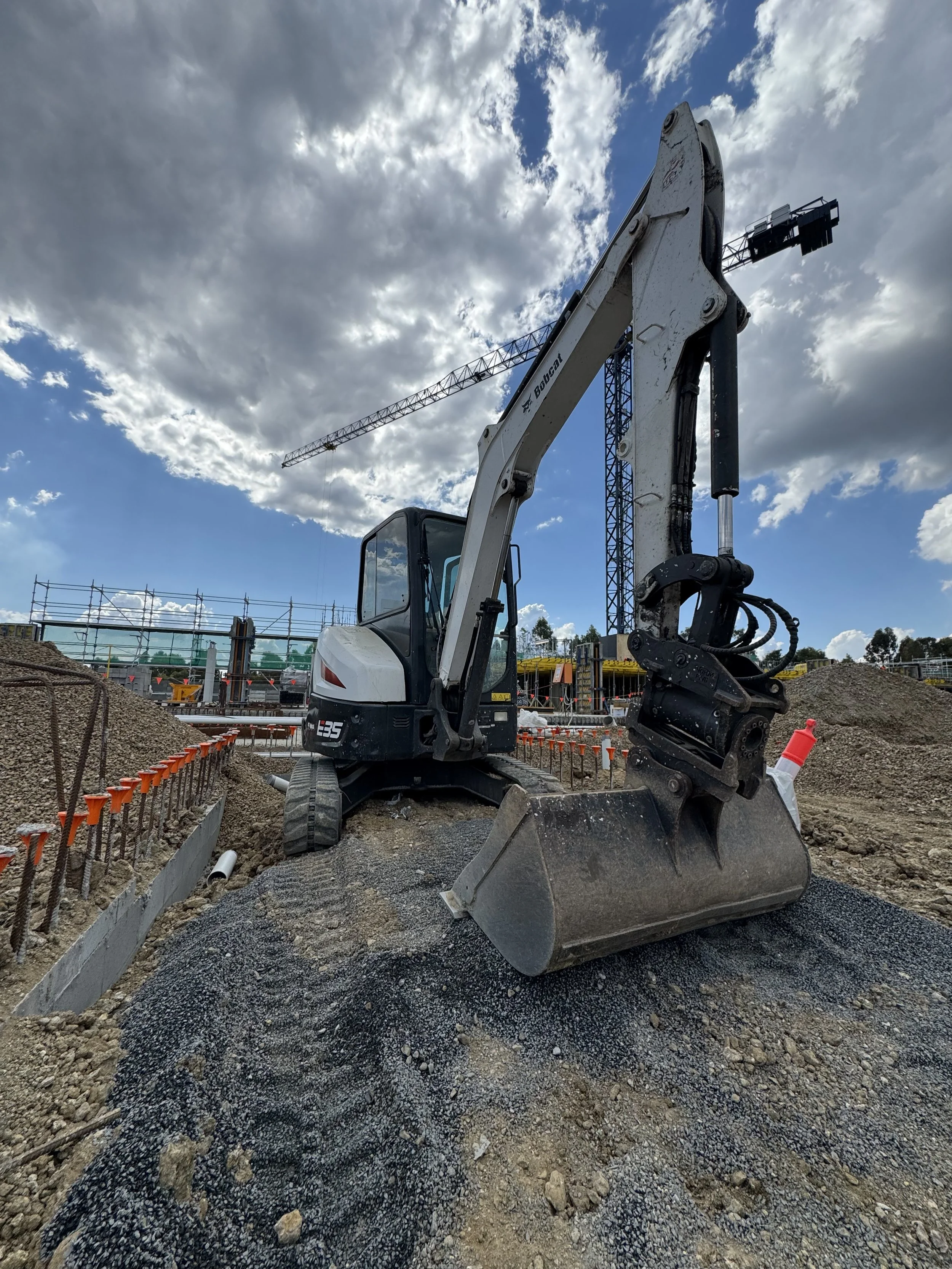 A construction site with a small excavator and a tower crane underneath a partly cloudy sky.