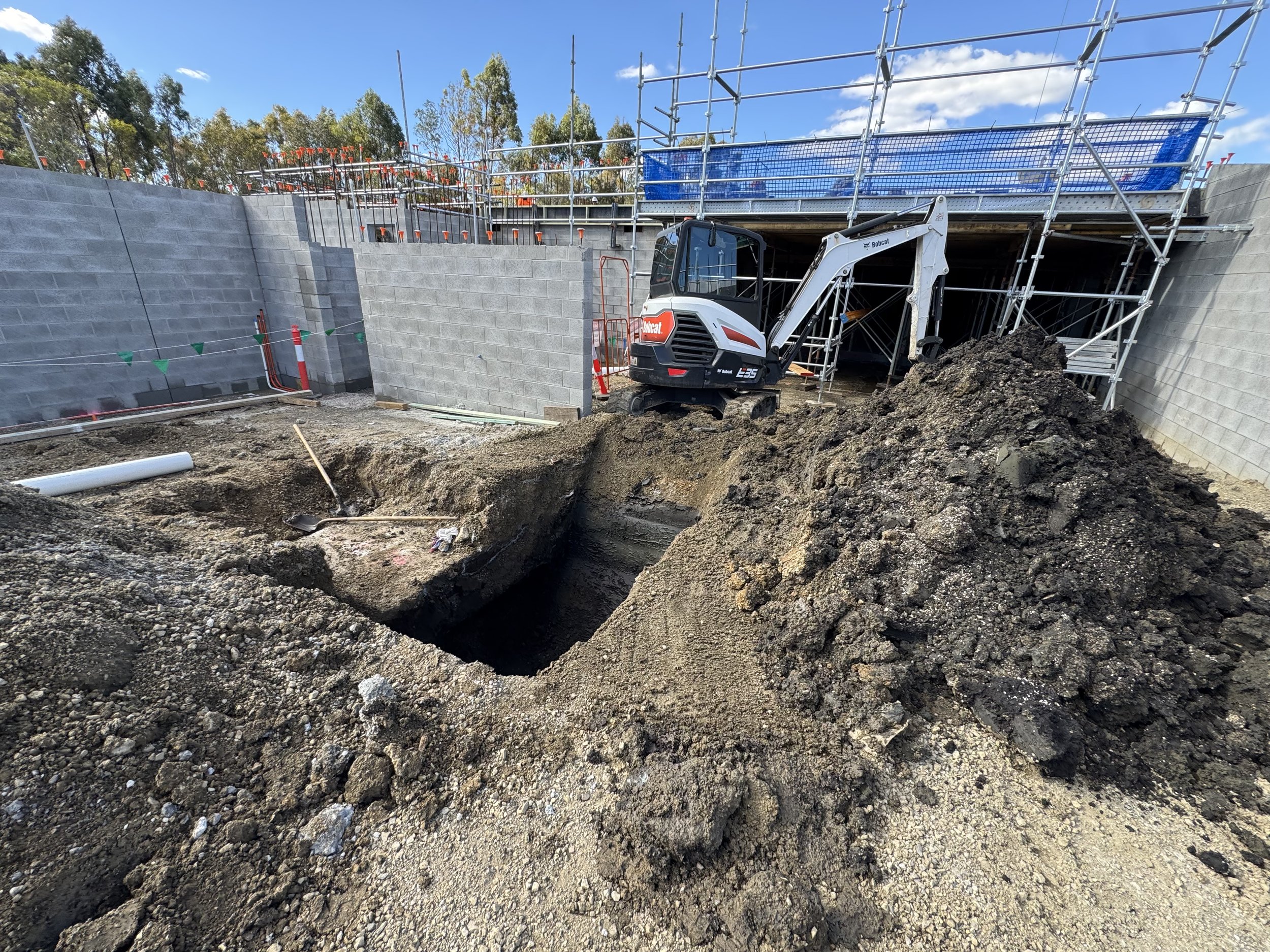 Construction site with a mini excavator digging a hole, scaffolding and cinder block walls, and a blue sky with clouds.
