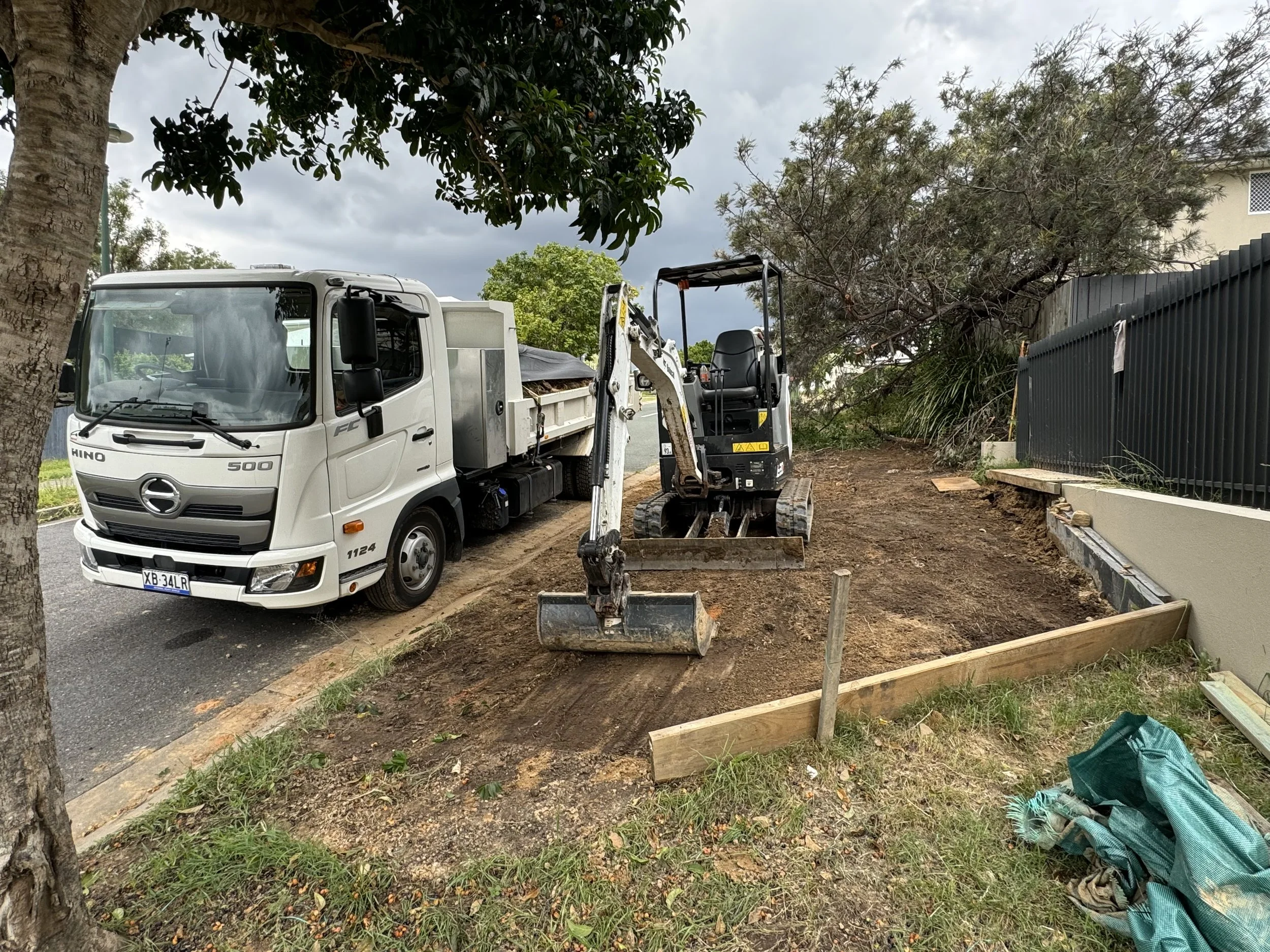 A construction site with a white truck carrying soil and a small excavator on uneven ground, surrounded by trees and a fence, under cloudy skies.