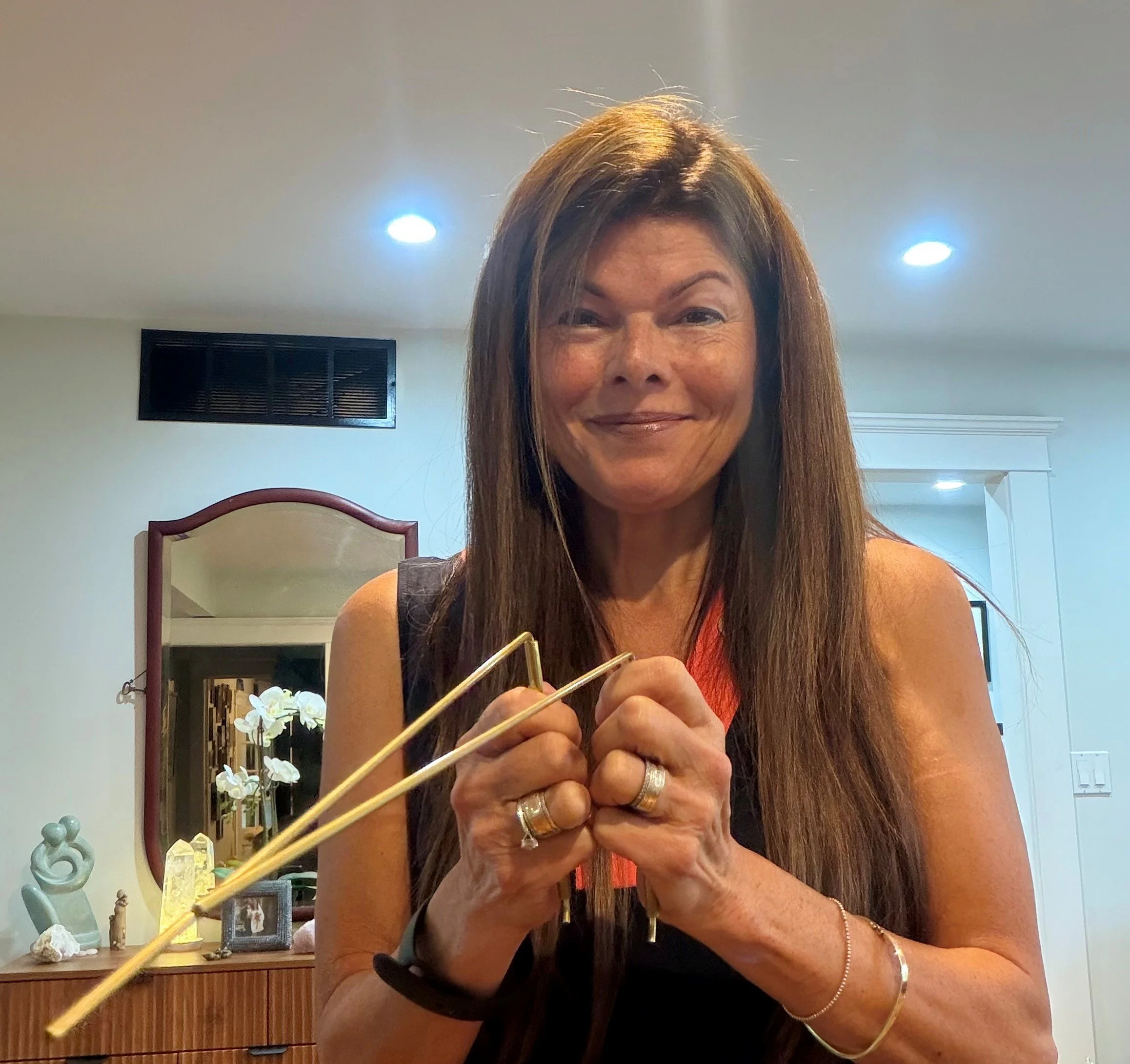 Tina holding dowsing rods indoors, with a background of a cabinet with decorative items and a mirror.