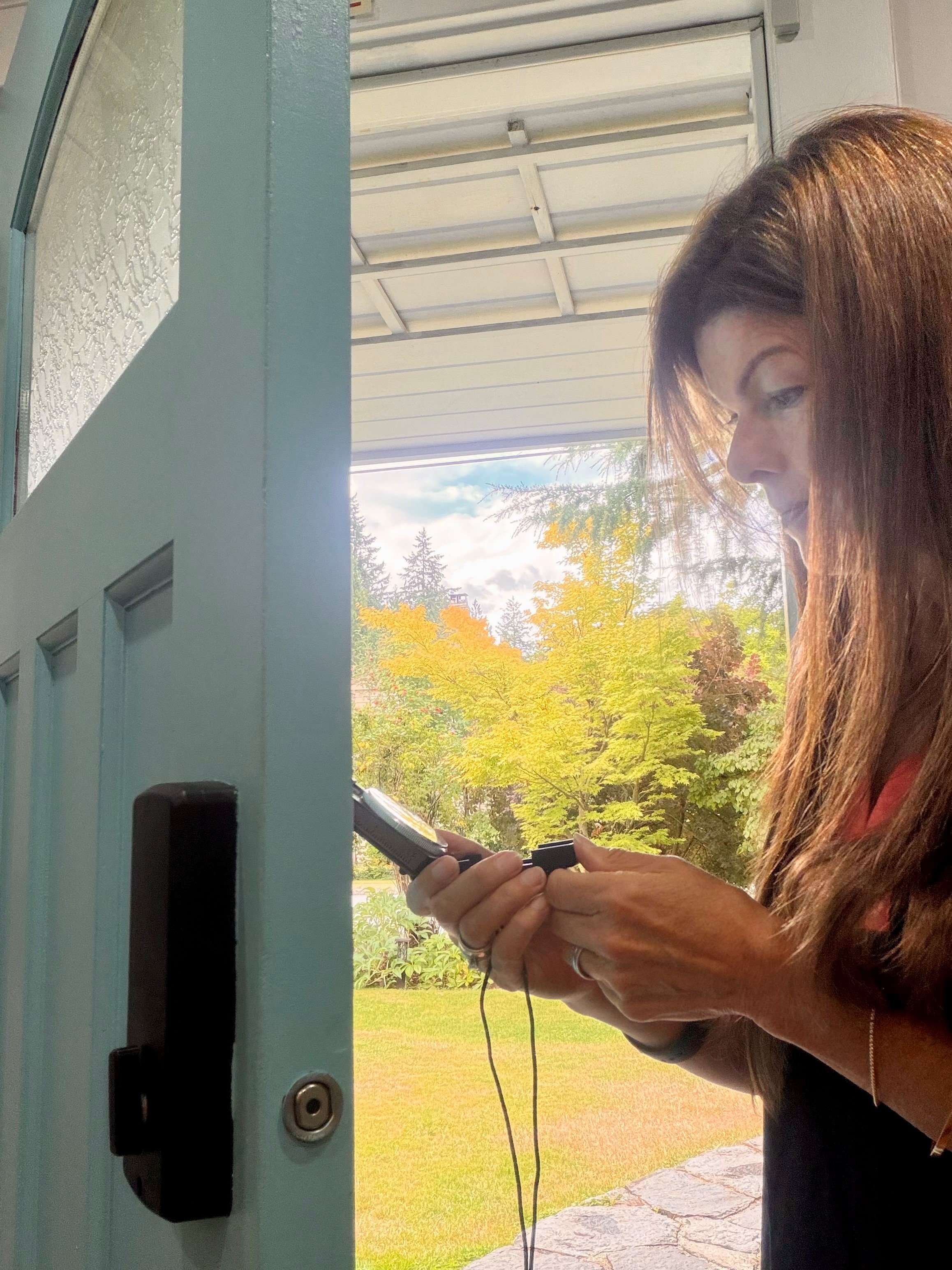 Tina looking at her compass, standing outside near a building with a green lawn and trees with colorful autumn leaves.