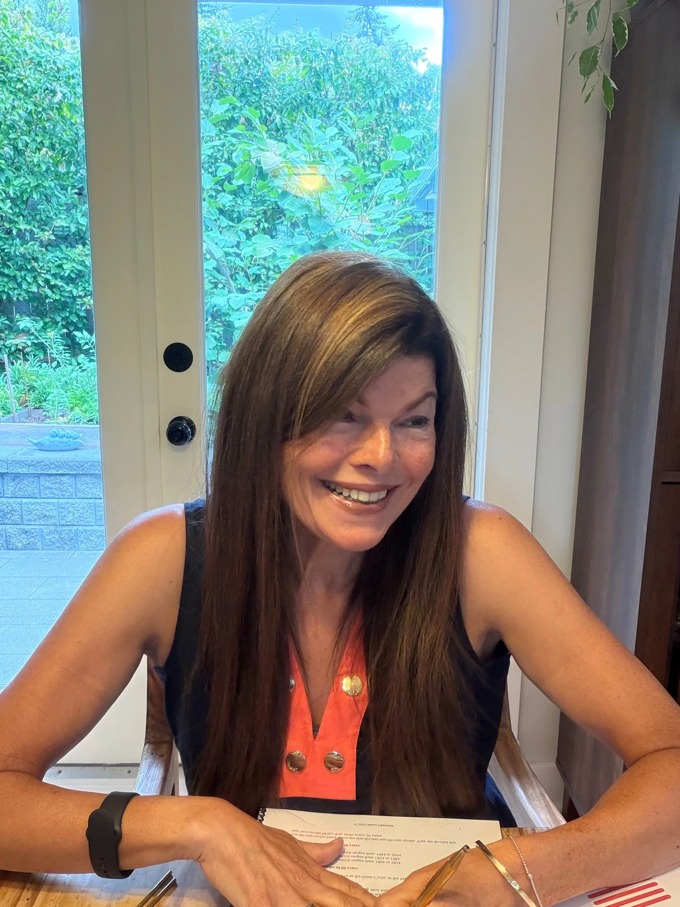 Tina sitting at a wooden table near a window with green foliage outside. She appears to be signing a document with a pen, wearing a black watch, a bracelet, and a sleeveless top with gold buttons.