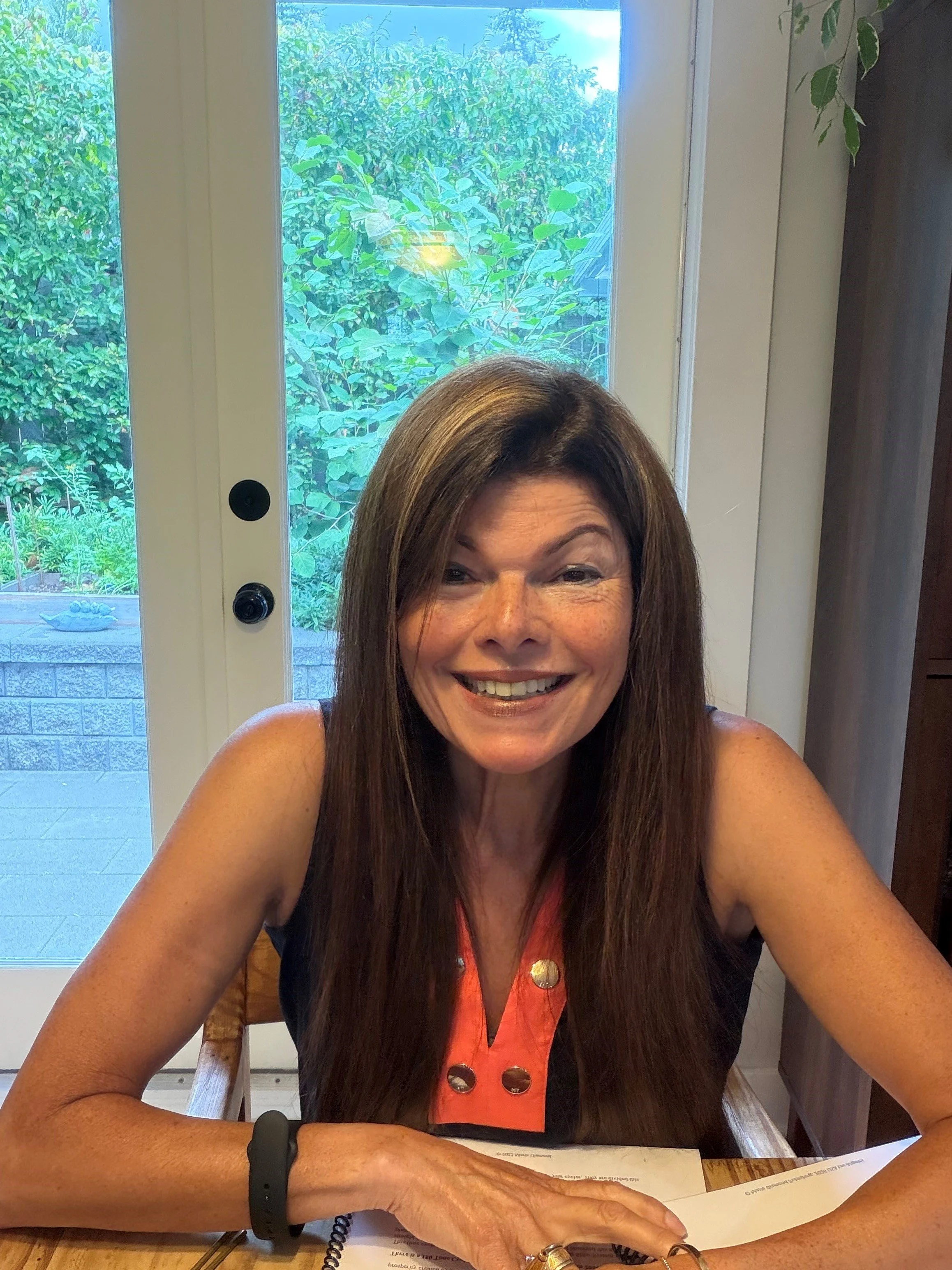 Tina smiling, sitting at a table with a menu, in front of a glass door with a view of greenery outside.