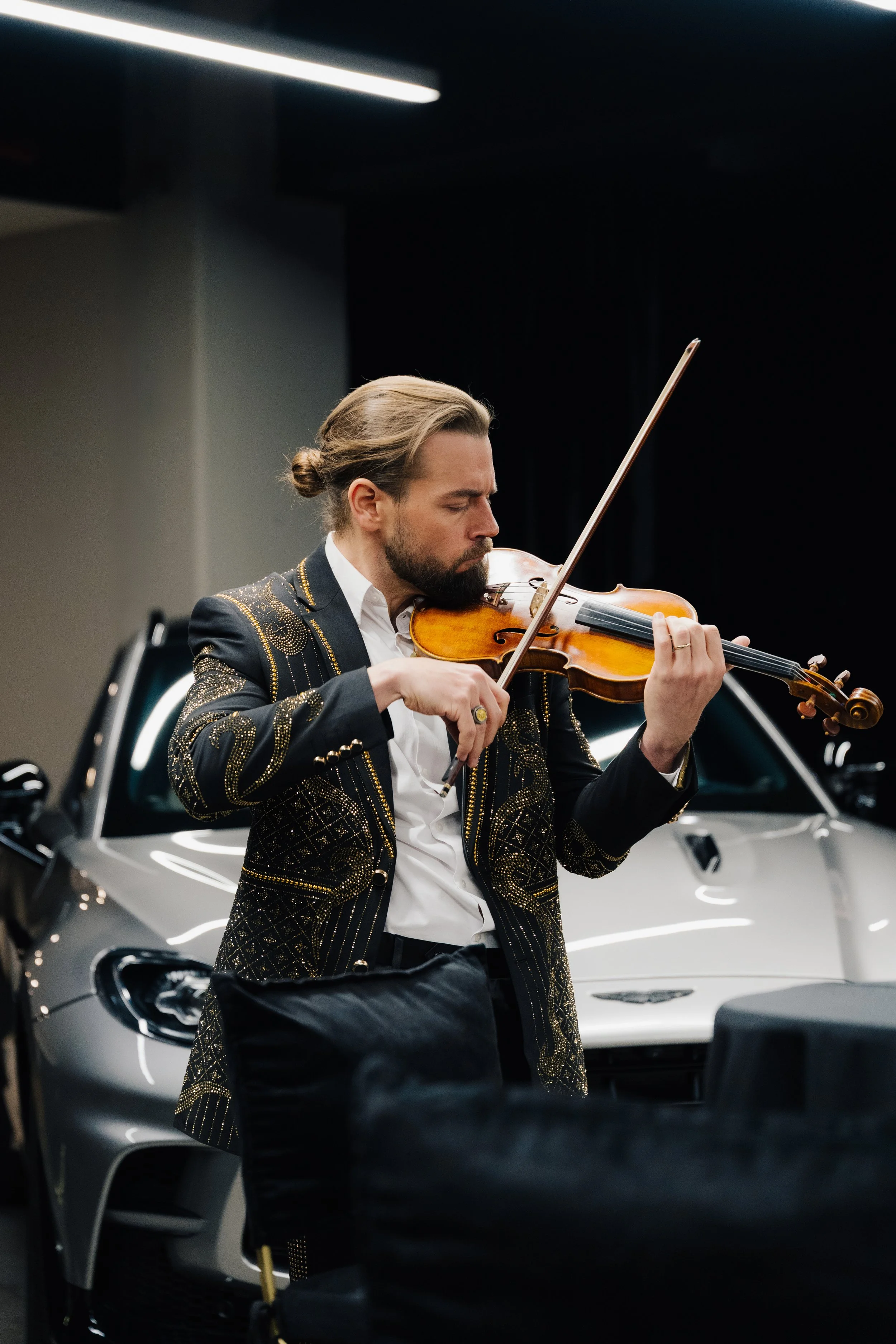 Markus, hombre tocando violín en un evento, con chaqueta negra con detalles dorados, con autos de lujo al fondo, un Aston Martin Valhala, en un entorno interior en la CDMX