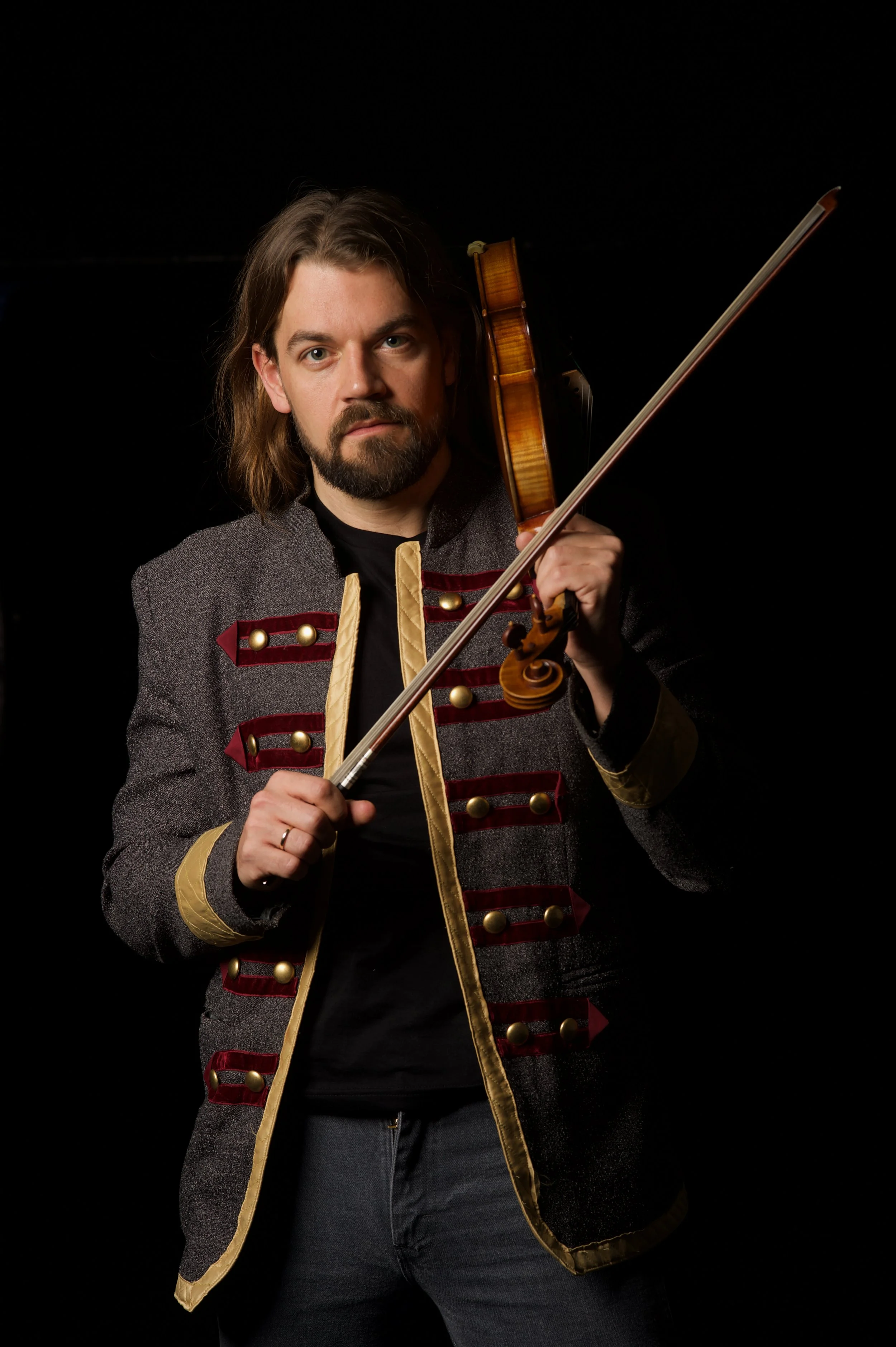 Hombre con barba y cabello largo, vistiendo chaqueta de estilo militar, sosteniendo un violín y un arco, con fondo negro.