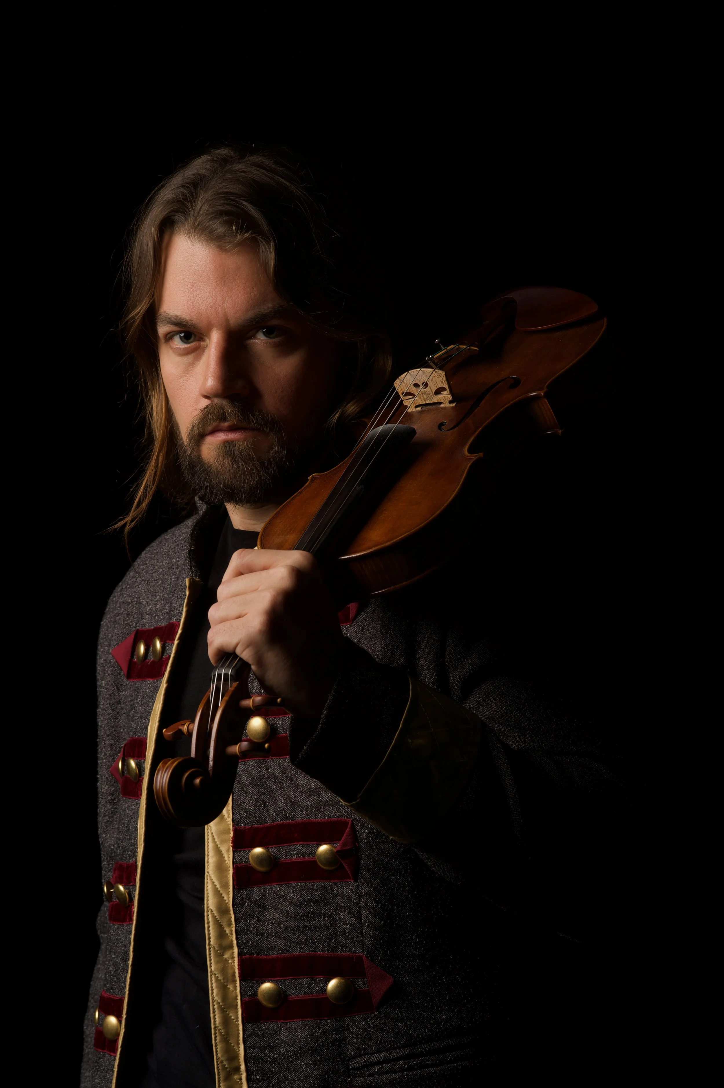 Hombre con barba y cabello largo sosteniendo un violín, vestido con chaqueta gris con detalles en dorado y rojo oscuro, fondo negro.