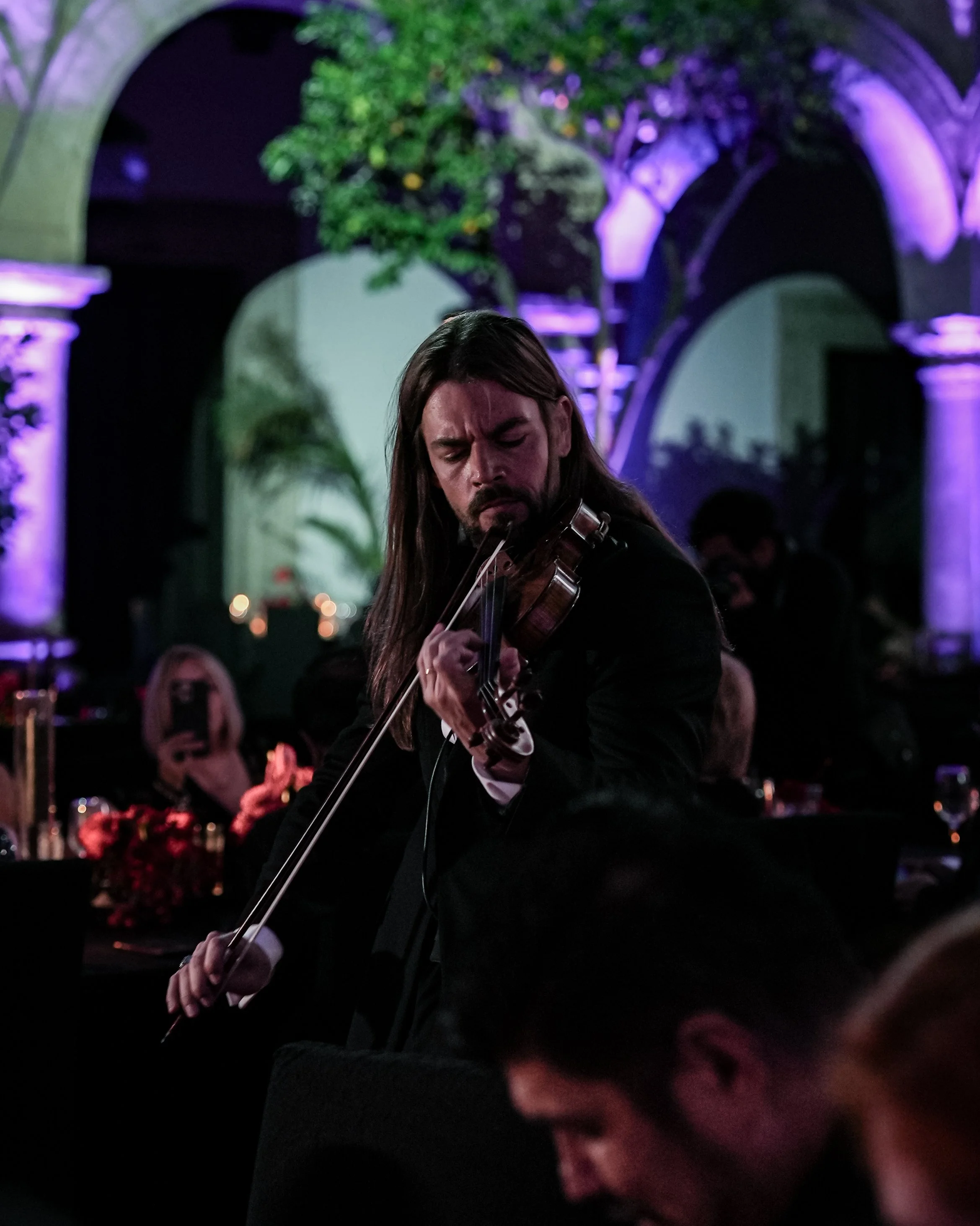 Hombre tocando violín en un escenario con fondo de arcos y luces moradas, en un ambiente elegante y oscuro. Gala Miss Universo Guadalajara. Palacio de Gobierno. Markus Violin.