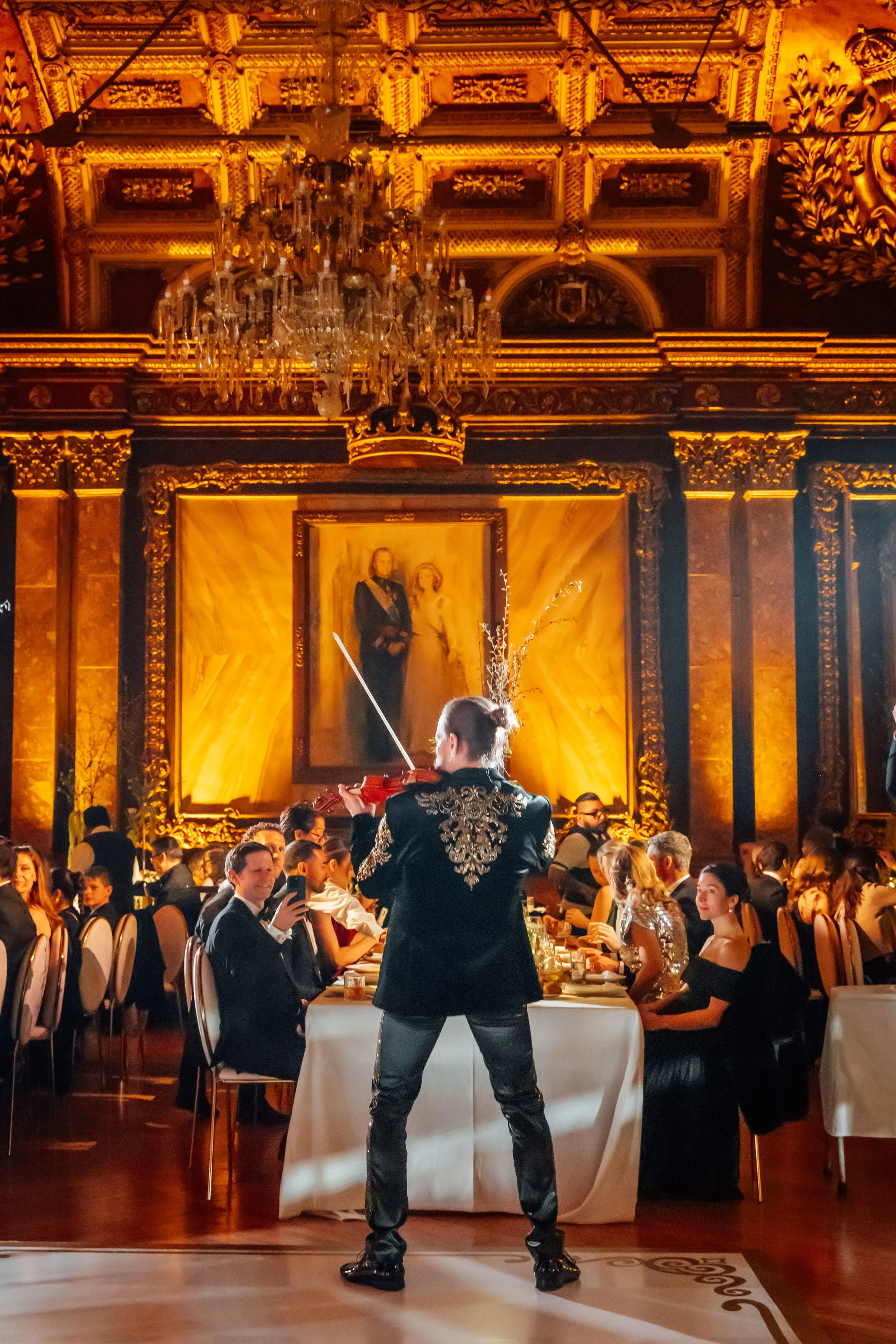 Hombre tocando violín en una cena elegante en un salón decorado con oro y una pintura de fondo, con muchas personas sentadas en mesas. La sala tiene un candelabro y un techo elaborado.