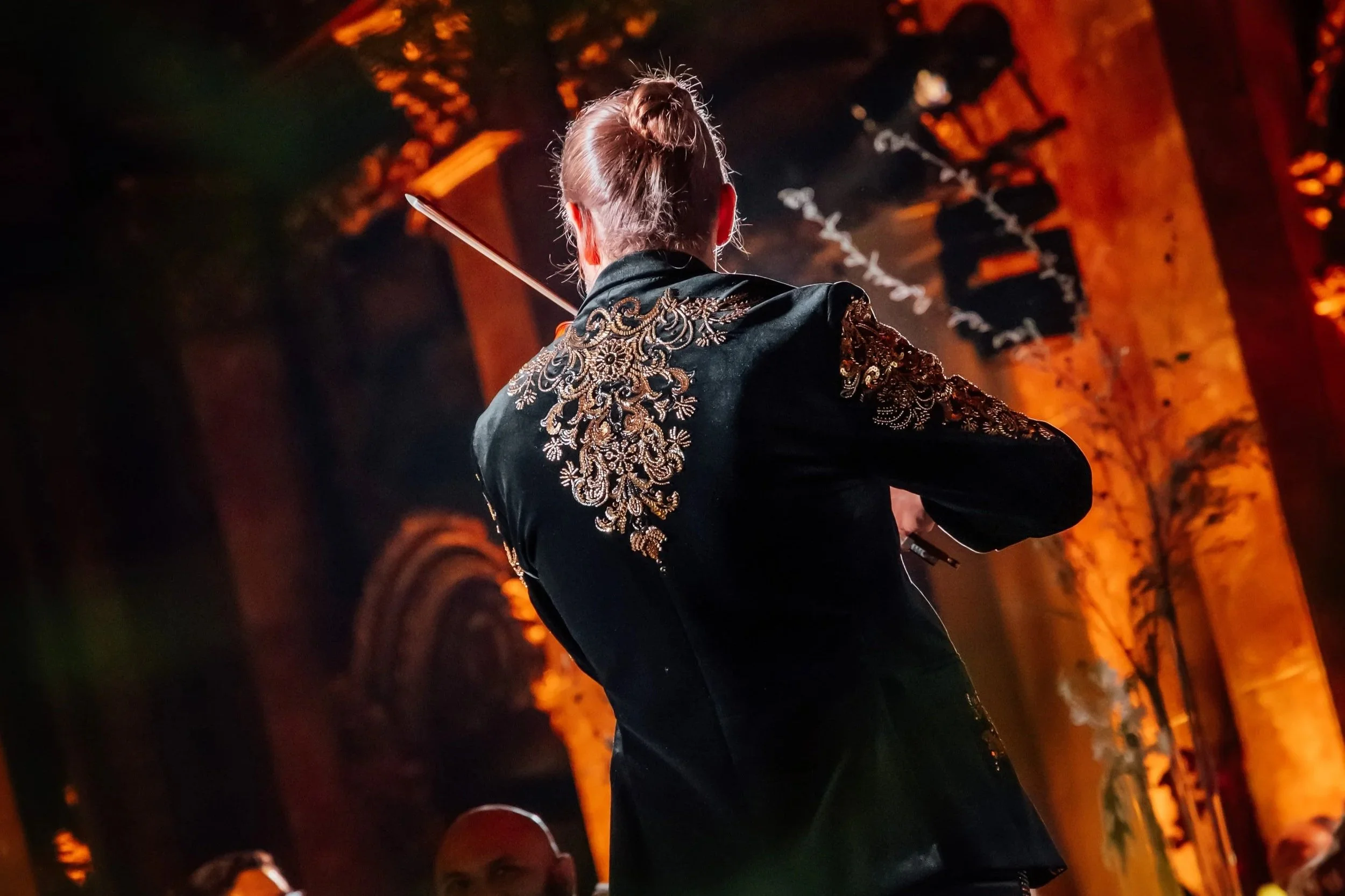 Markus vista de espaldas, tocando violín en un escenario iluminado con focos anaranjados, con detalles bordados dorados en la vestimenta. CDMX, Casino Español.