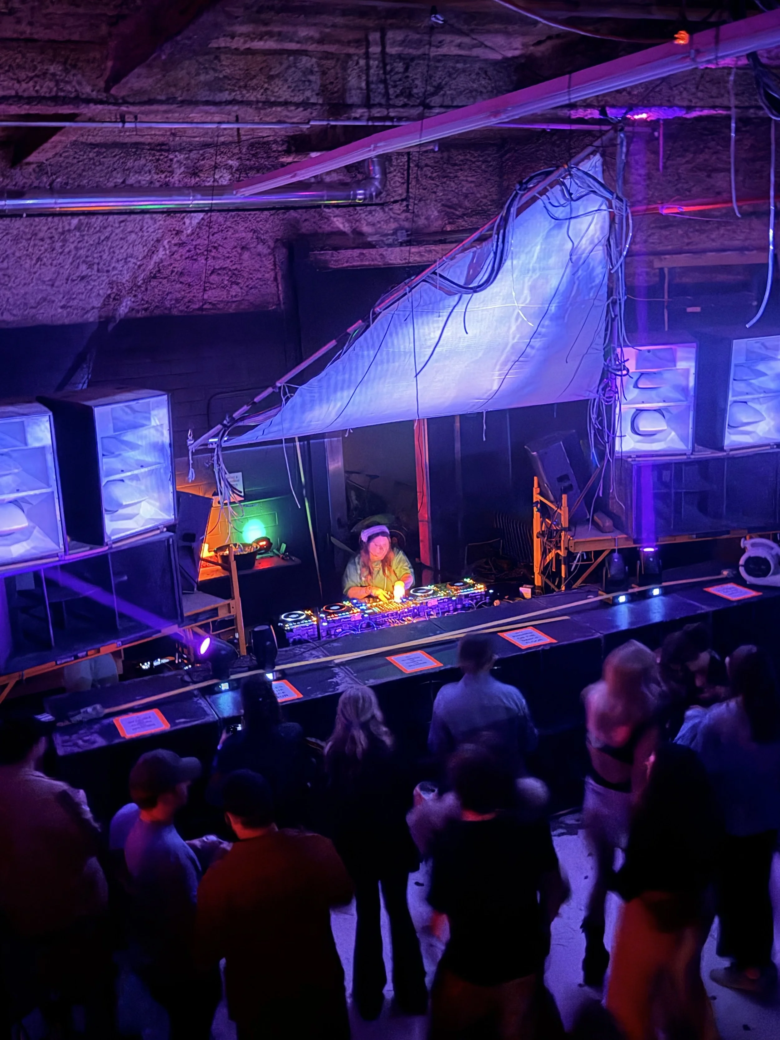 A DJ performs at a nightclub with an audience watching. The DJ is using a turntable setup under colorful stage lighting, with a rock wall backdrop and people standing in front.