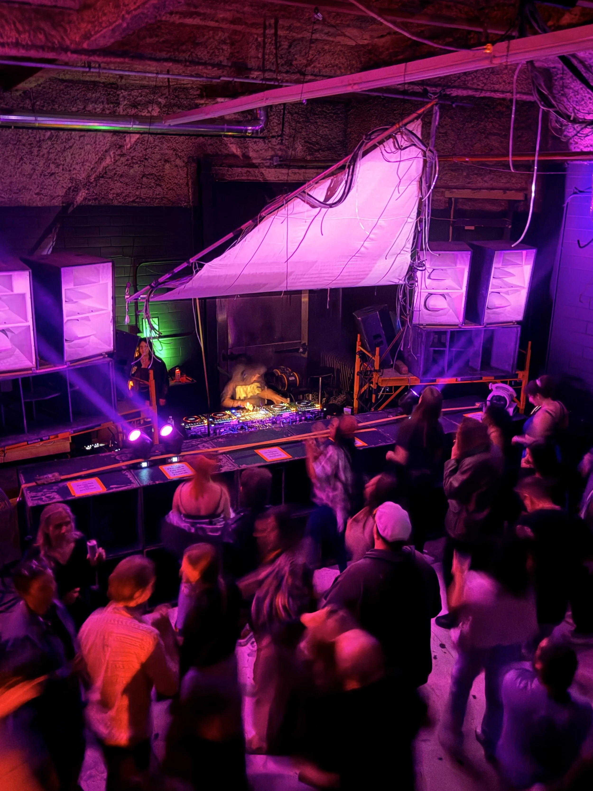 People gathered in a dimly lit venue with purple and pink lighting, DJ equipment on a stage, and an industrial ceiling with exposed pipelines.