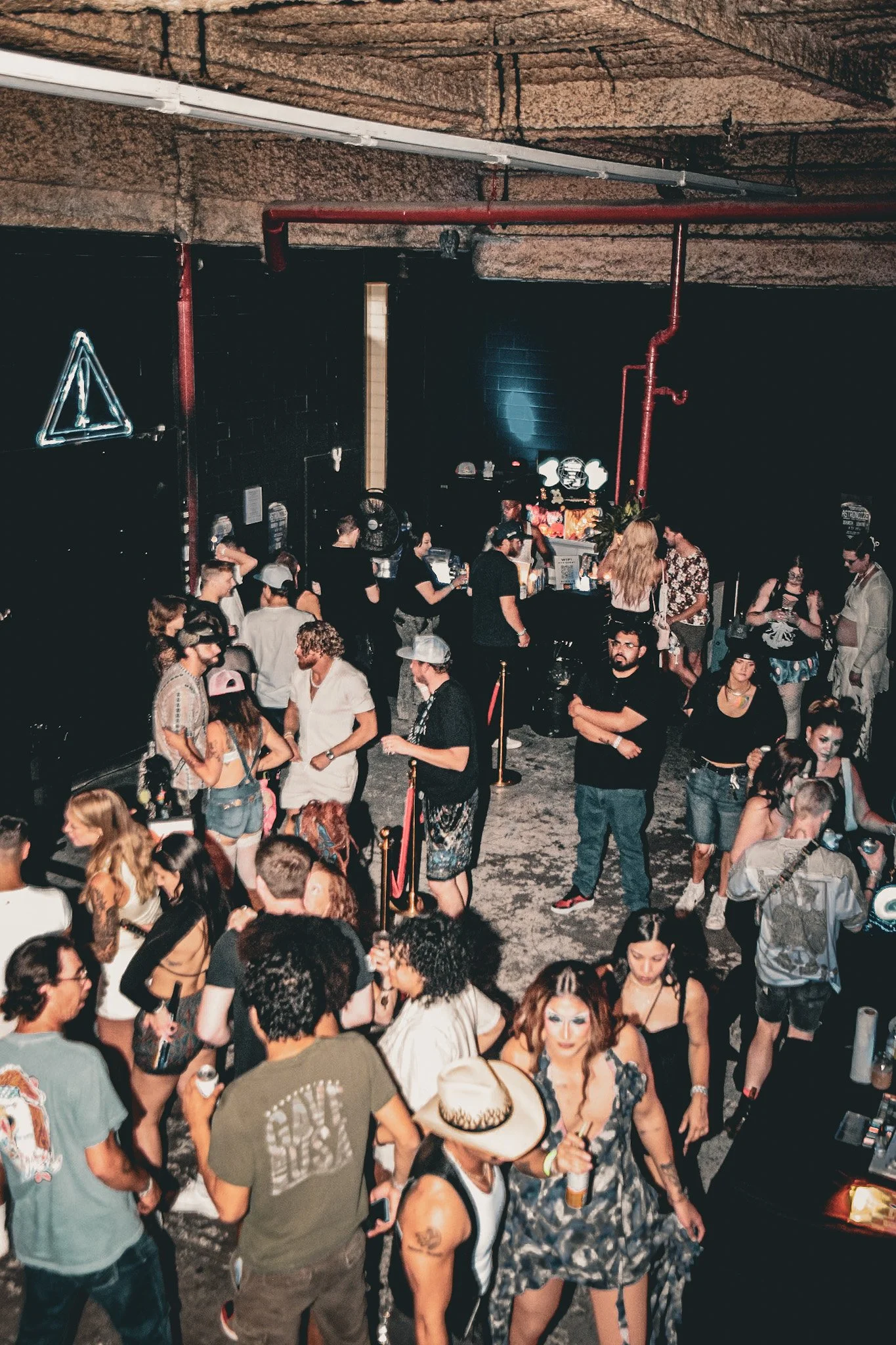 A crowded indoor event with people socializing, some with tattoos, wearing casual and festival-style clothing, in a venue with a black wall, neon signs, and industrial ceiling features.