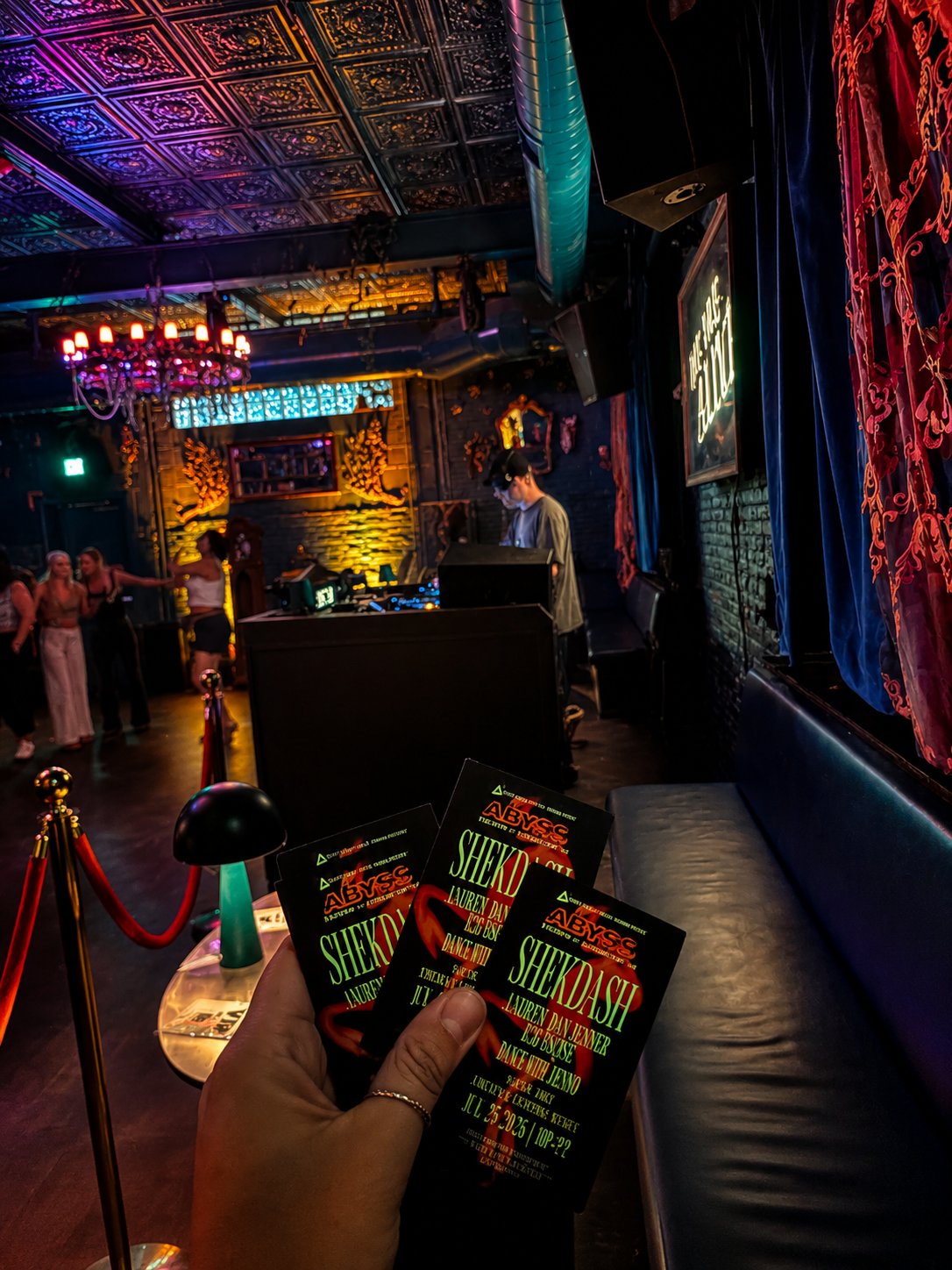 A hand holding three event flyers for a party titled 'Shekash' at Abyss nightclub, with a DJ performing on stage in the background, and a few people dancing on the dance floor. The club's interior features ornate ceiling tiles, colorful lighting, and velvet curtains.