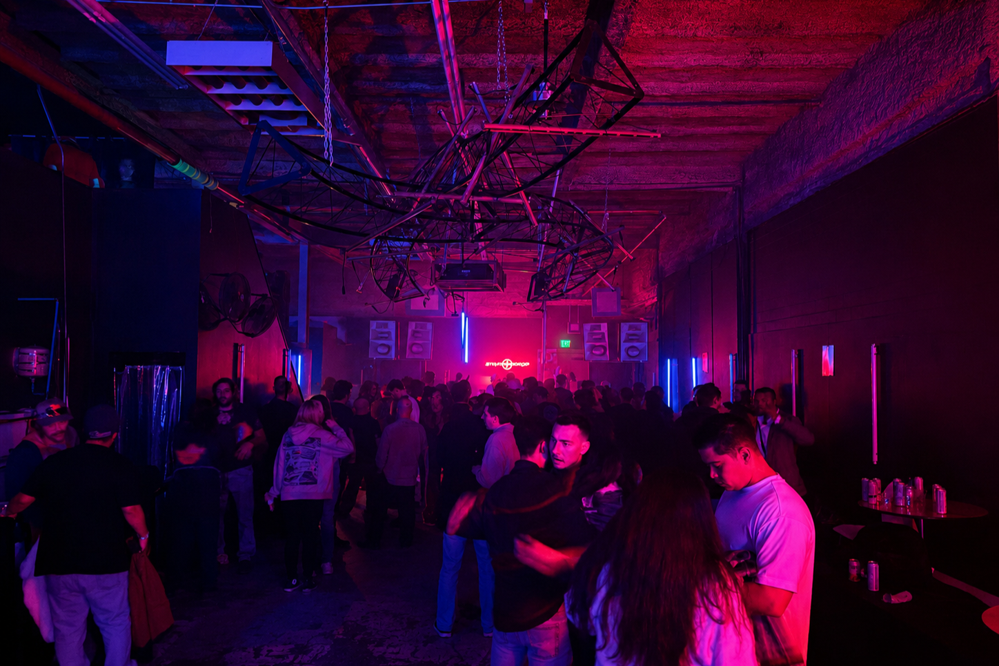People dancing and socializing in a dark nightclub with purple and pink neon lighting, industrial ceiling beams, and a DJ setup at the front.