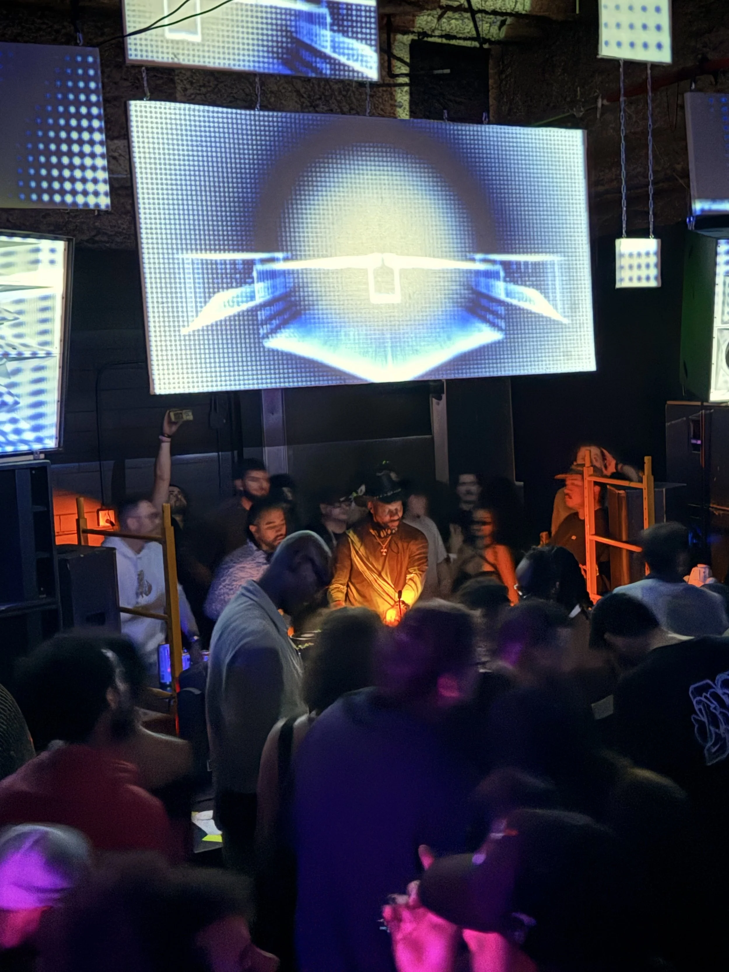 A digital art installation featuring multiple LED screens displaying blue and white pixelated graphics in a dark room with a crowd of people watching.
