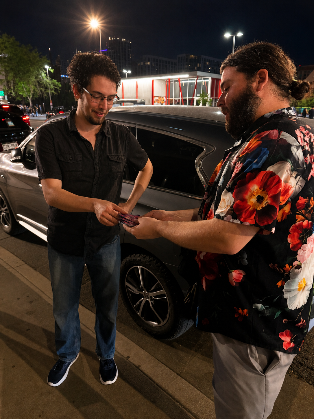 Two men exchanging items on a city street at night, one wearing a black shirt and jeans, the other in a colorful floral shirt, with parked cars and illuminated buildings in the background.