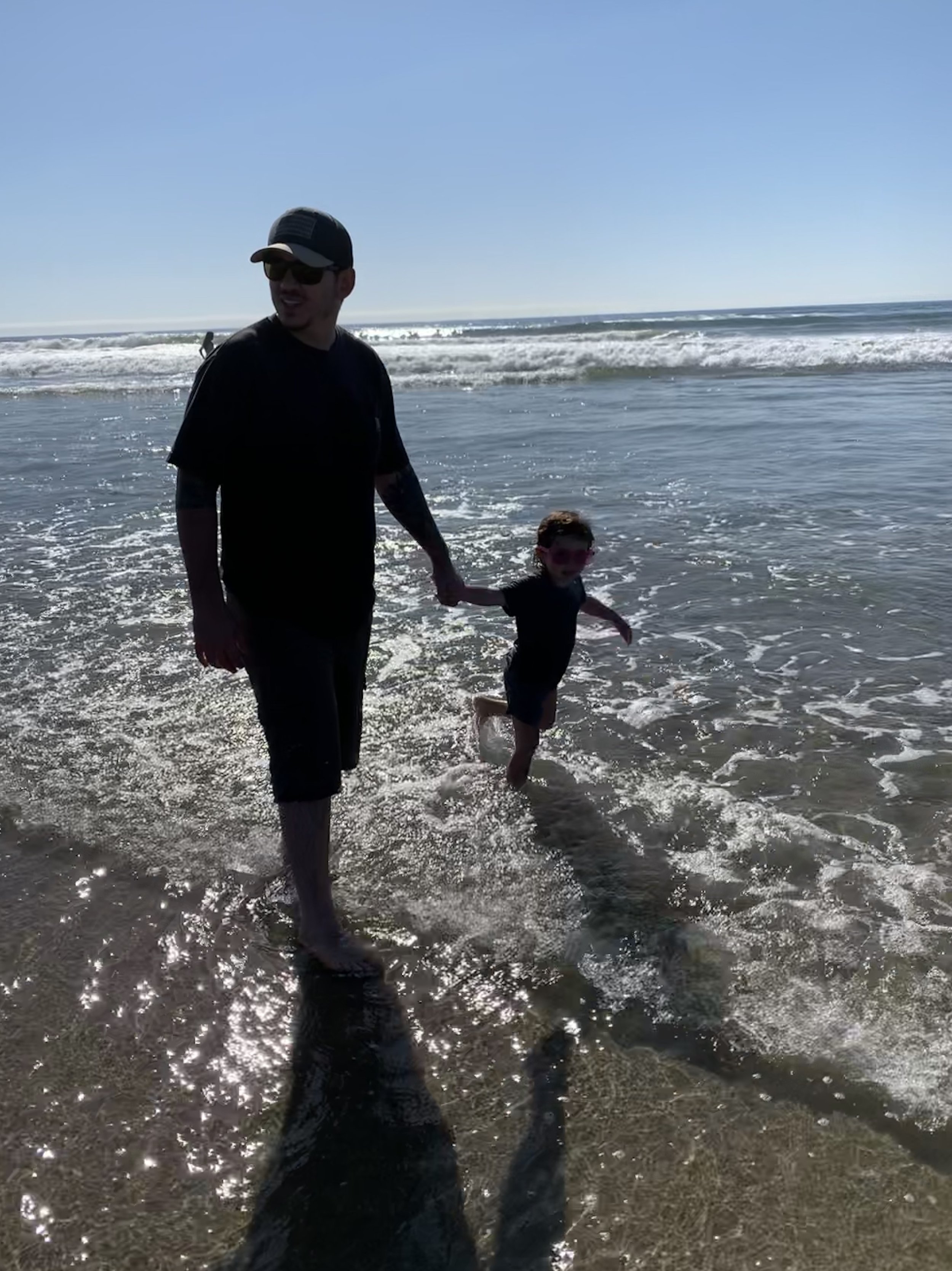 A man and a young boy holding hands and walking in the shallow water of a sunny beach with waves in the background.