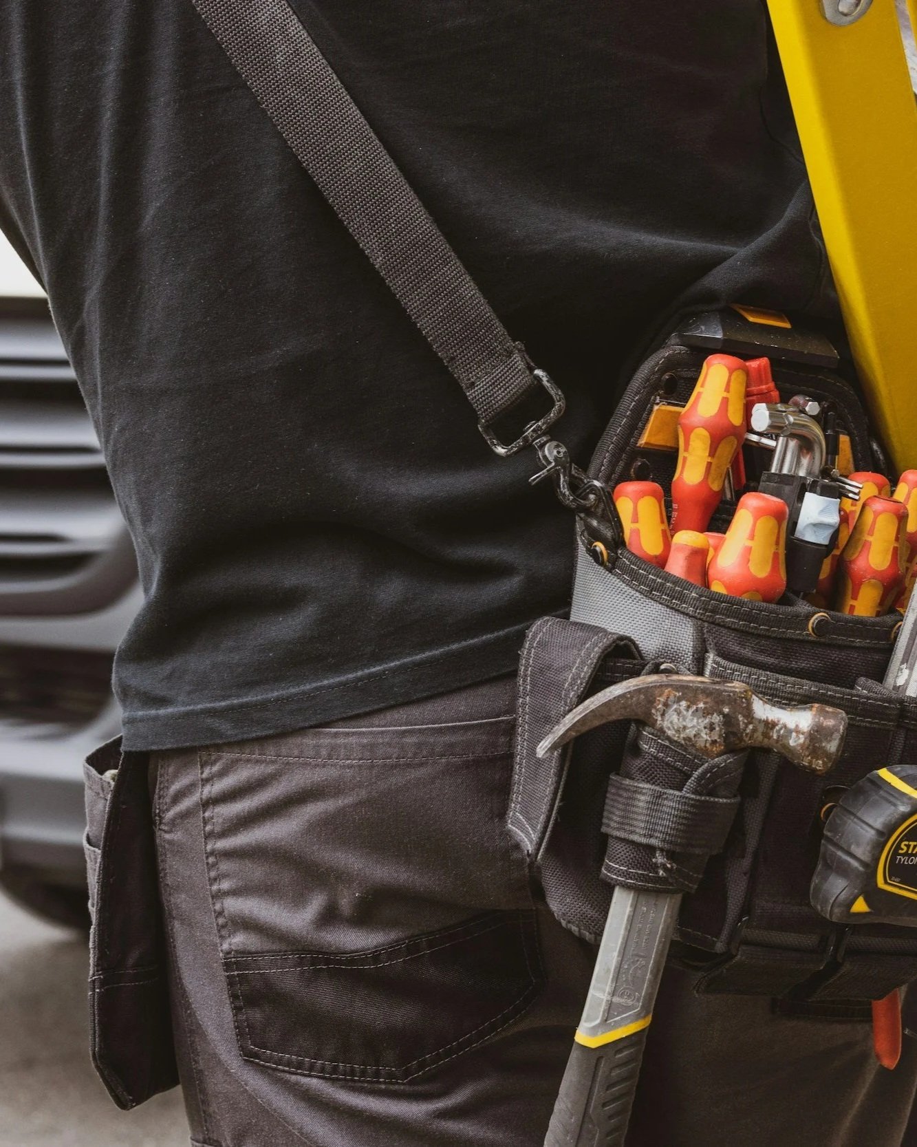 A worker wearing a black shirt and black pants has a tool pouch with screwdrivers, a hammer, and other tools attached to their waist, with a yellow and black power drill next to them.