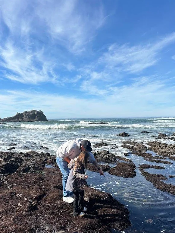 A man and a young girl exploring tide pools on a rocky beach with the ocean and blue sky in the background.