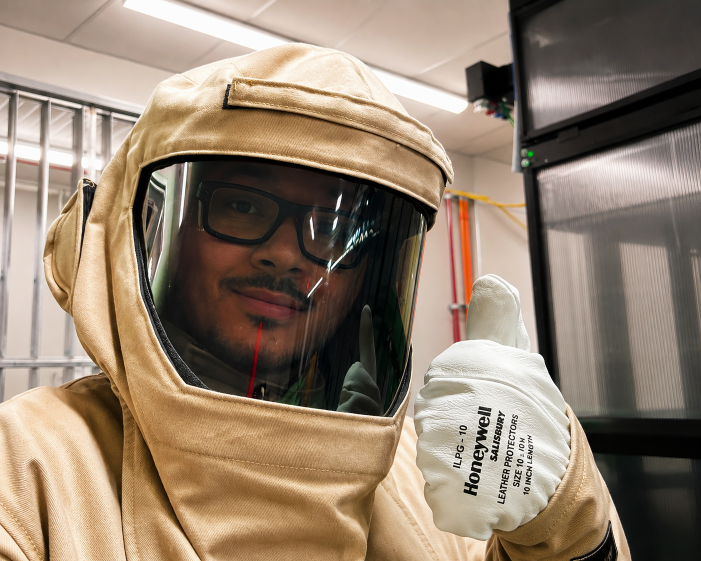 A man in protective welding gear, including a beige hood and a face shield, giving a thumbs-up while wearing white gloves with black text.