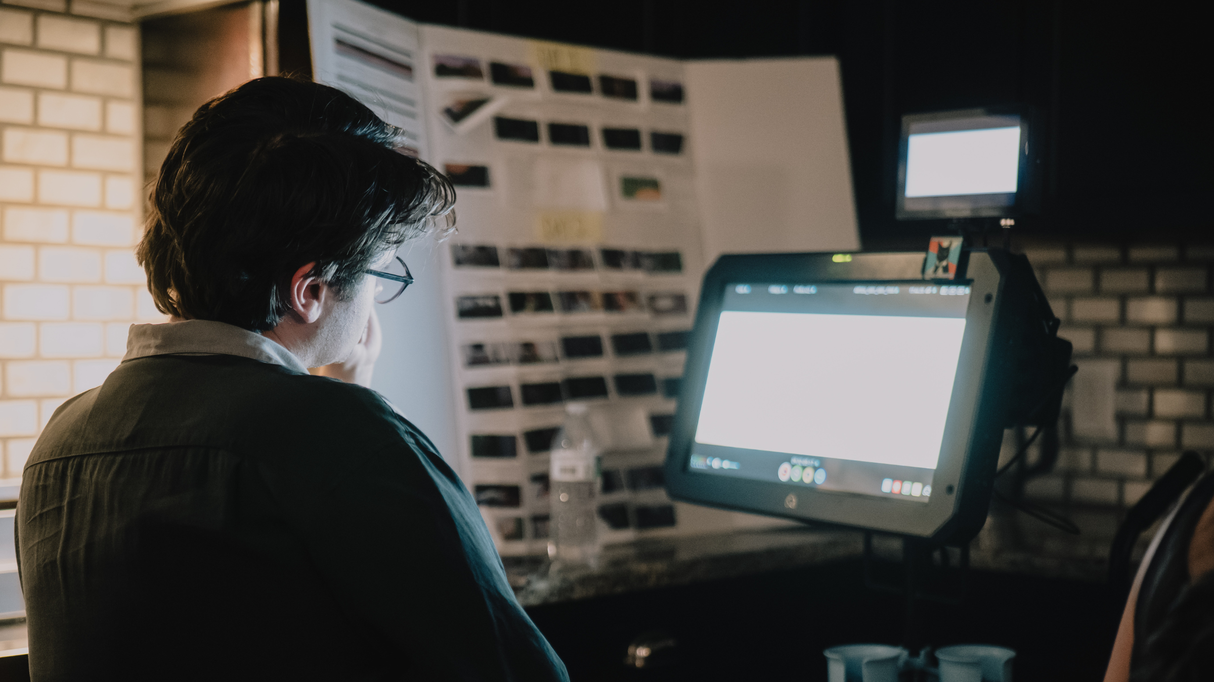 A person wearing glasses is seated at a desk in front of a computer monitor, with a large whiteboard filled with photographs or images behind them, in a dimly lit room.