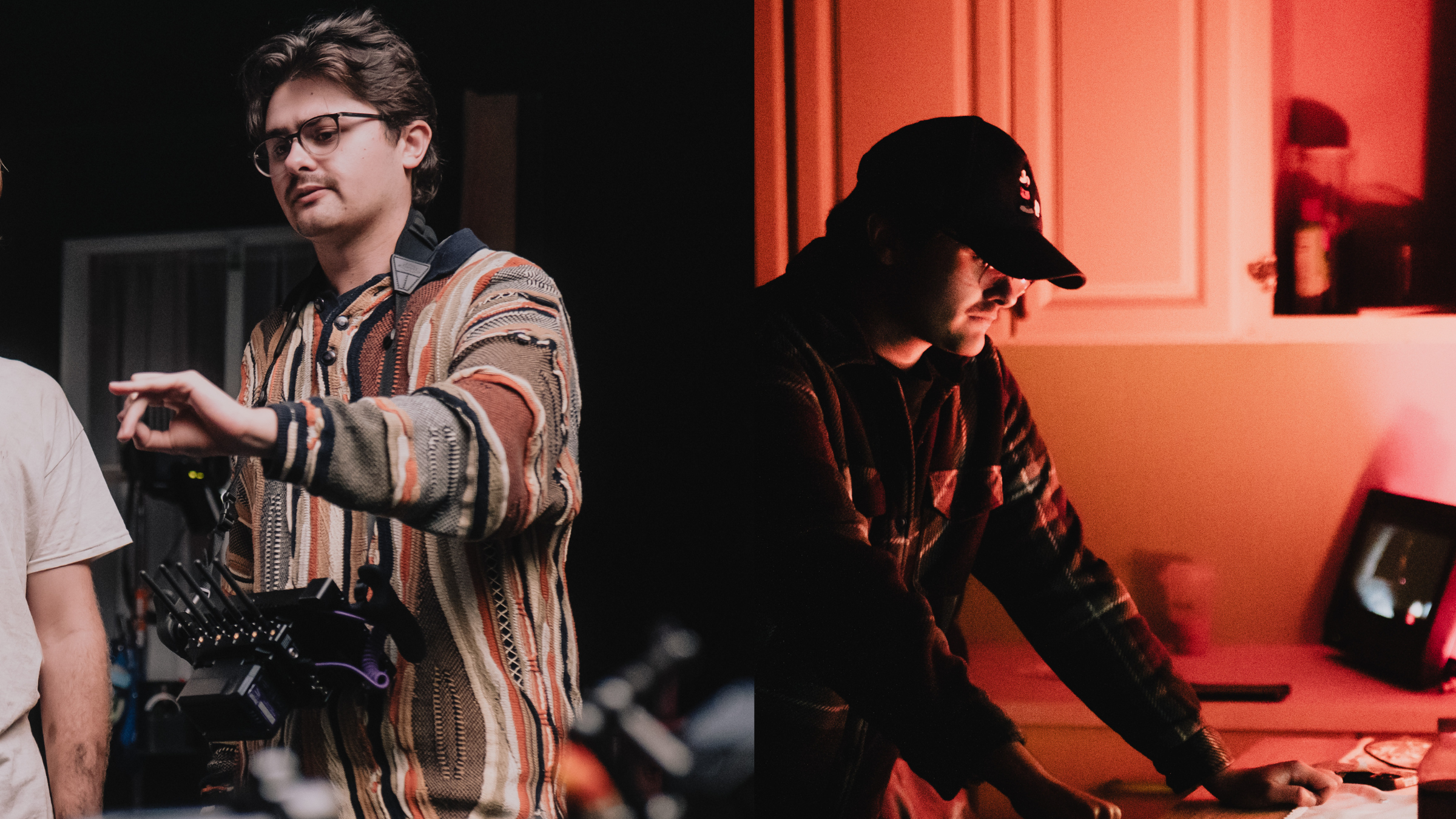 A young man with glasses and a patterned sweater is standing in a dimly lit setting, possibly a film set or studio, with equipment around him. To his right, another person wearing a cap and a plaid jacket is focused on working at a computer with red ambient lighting.