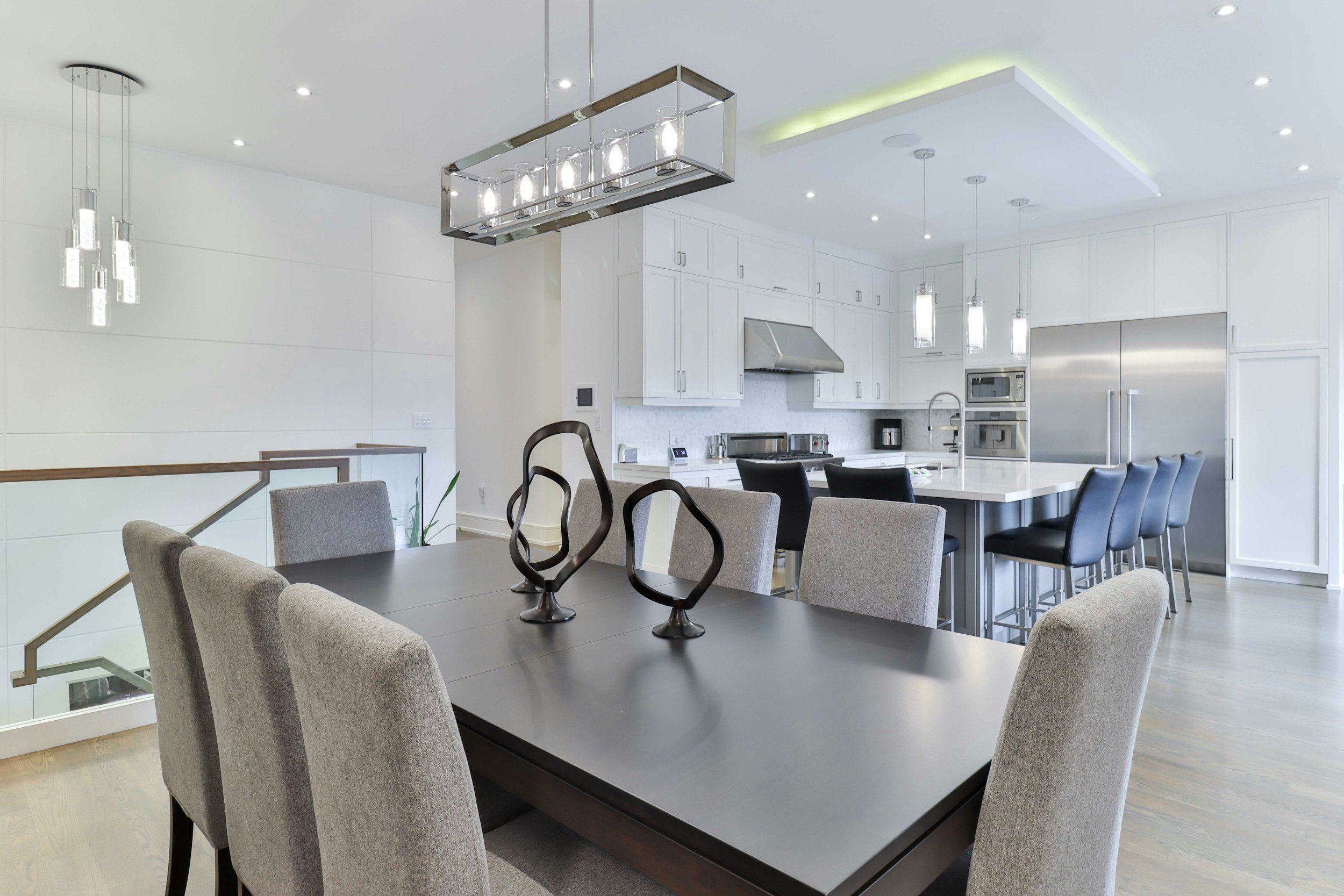 Modern kitchen and dining area with white cabinetry, stainless steel appliances, black dining table, and upholstered chairs, illuminated by stylish pendant and ceiling lights.