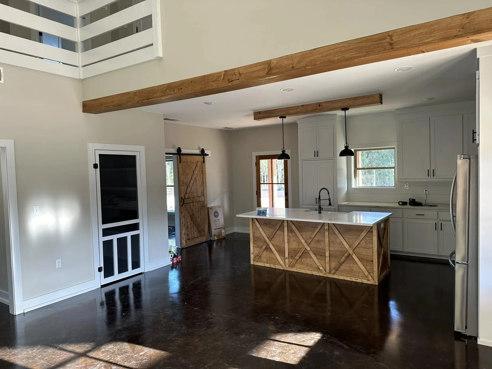 Interior view of a modern kitchen with white cabinets, a kitchen island with a wooden base, pendant lighting, and wooden beams on the ceiling. Large windows let in natural light, and there is a sliding barn door.