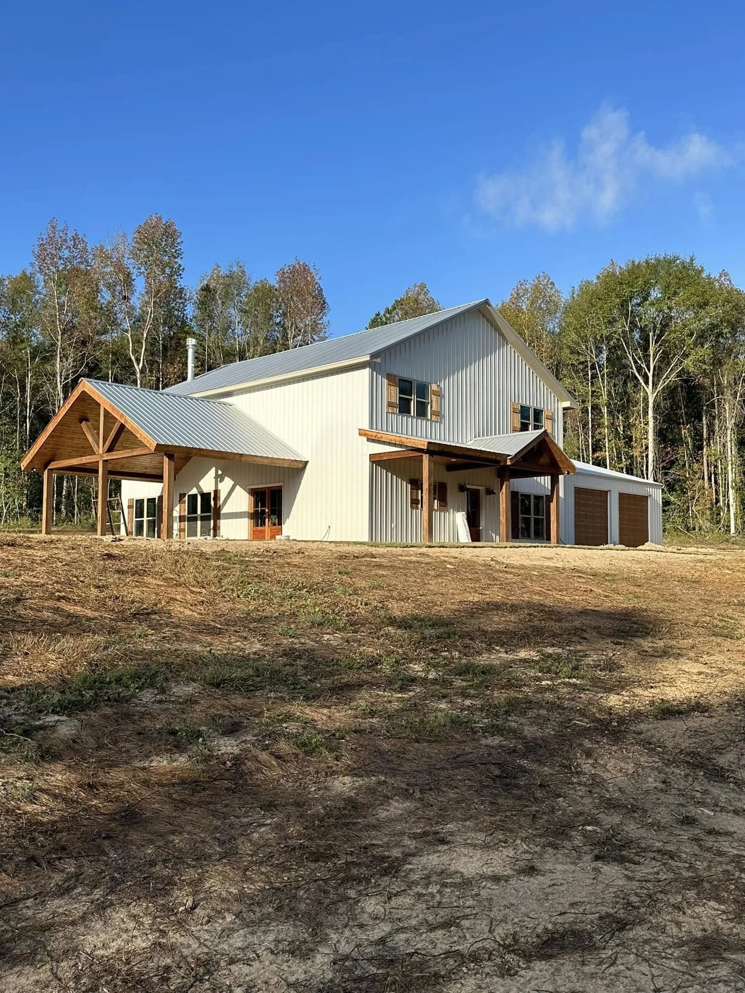 A house with a metal roof, white siding, and brown wooden accents, situated on a cleared patch of ground with a forest in the background and a partly cloudy sky overhead.
